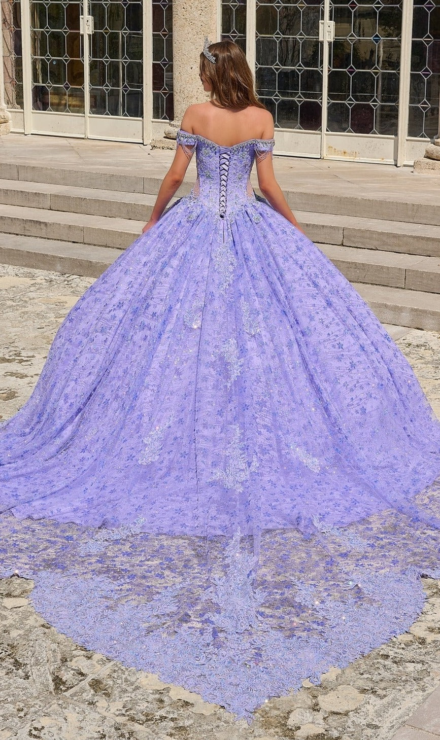 A woman stands outdoors in front of stairs wearing the Quinceanera Dress 54220 By Amarra, an off-the-shoulder lavender ball gown with a corset-style back, shimmering floral glitter tulle, lace train, and flowing silhouette.
