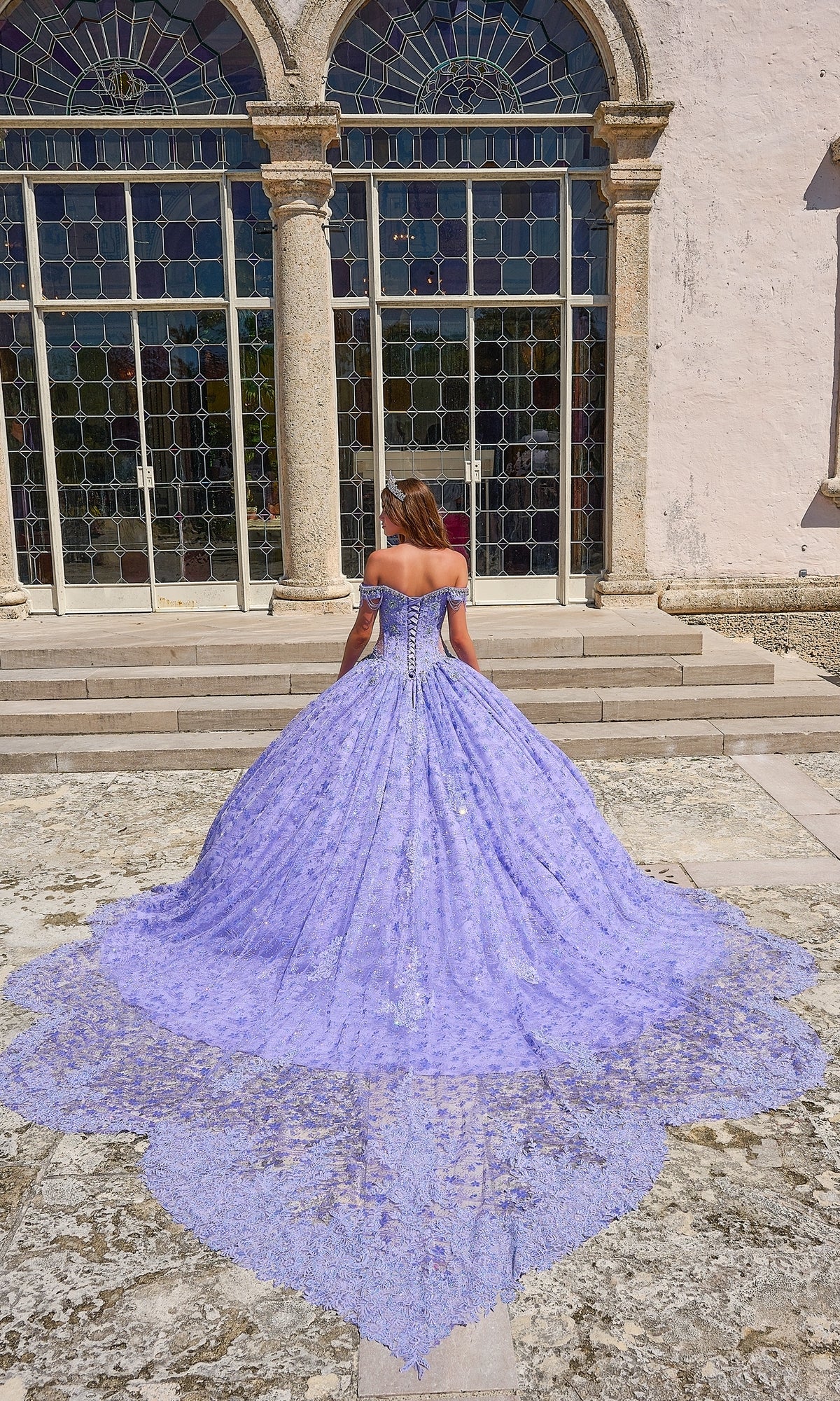 A woman in the Quinceanera Dress 54220 By Amarra, an off-the-shoulder lavender ball gown with a long, detailed train, stands on stone steps before glass doors and arched windows.