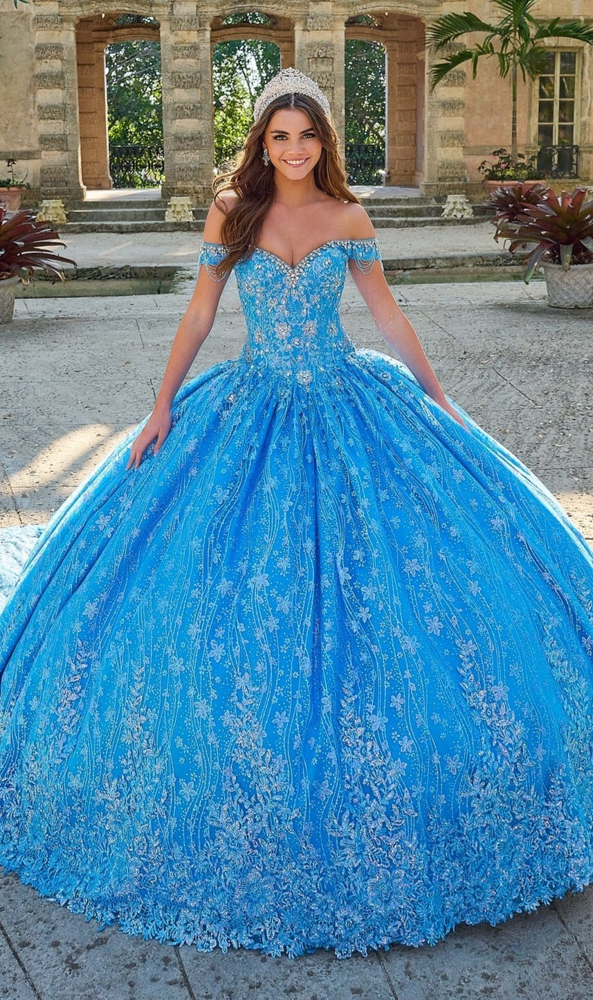 A woman in the Quinceanera Dress 54220 By Amarra, a sparkling blue off-the-shoulder ball gown with floral glitter tulle and a tiara, stands outdoors before an old stone building.
