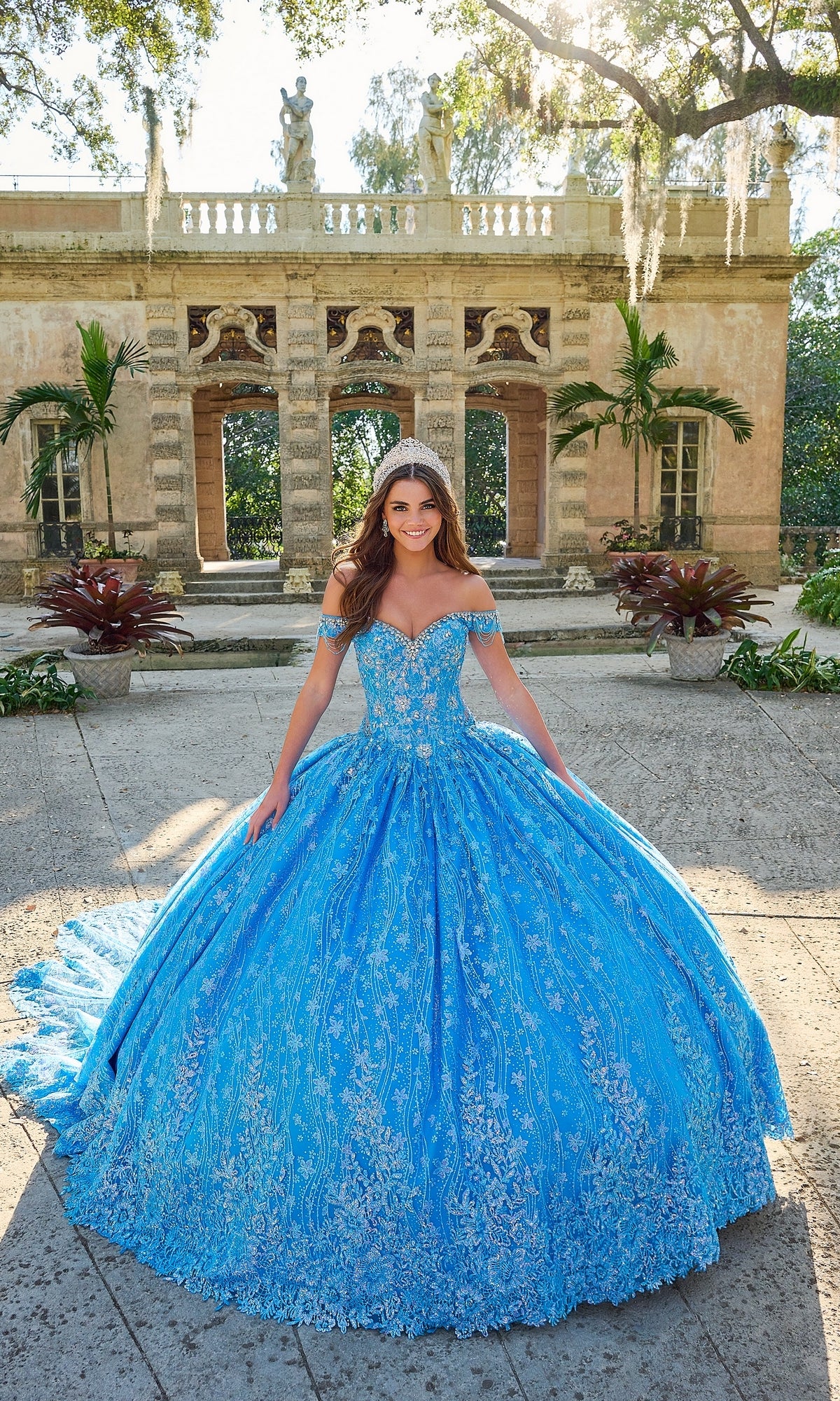 A woman in the Quinceanera Dress 54220 By Amarra, an ornate blue ball gown with off-the-shoulder sleeves, stands outdoors in front of a historic stone building with columns and greenery.