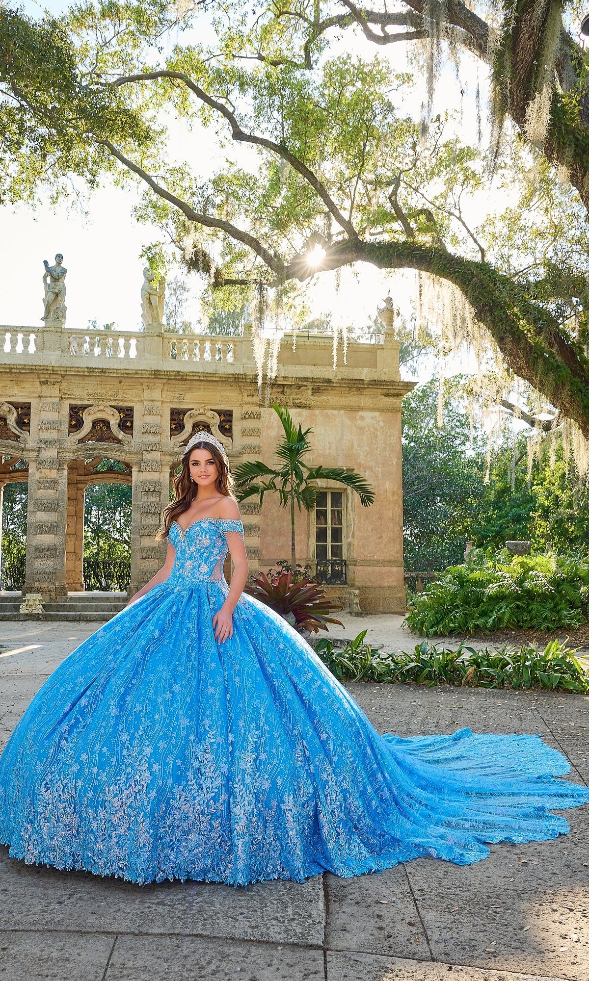 A young woman wears the Quinceanera Dress 54220 By Amarra, a bright blue off-the-shoulder ball gown with floral glitter tulle, standing outside an ornate stone building with arched columns and lush greenery.
