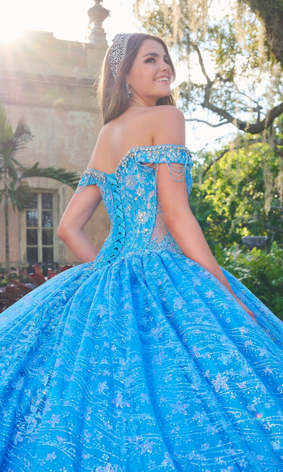 A young woman in the Quinceanera Dress 54220 By Amarra, featuring an ornate blue off-the-shoulder design and a shimmering headpiece, stands outside smiling over her shoulder.