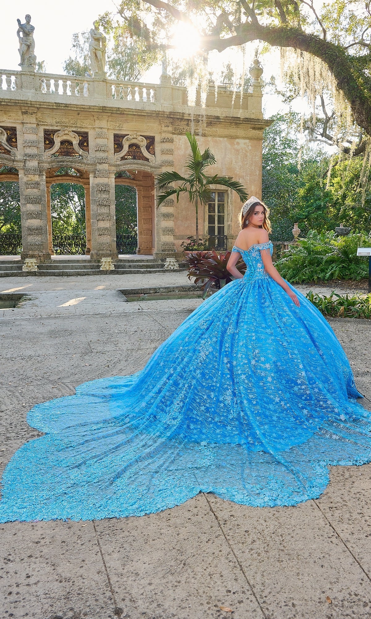 A woman stands outside a historic stone building in sunlight, wearing the Amarra Quinceanera Dress 54220—a blue, off-the-shoulder ball gown with floral glitter tulle and a long train.