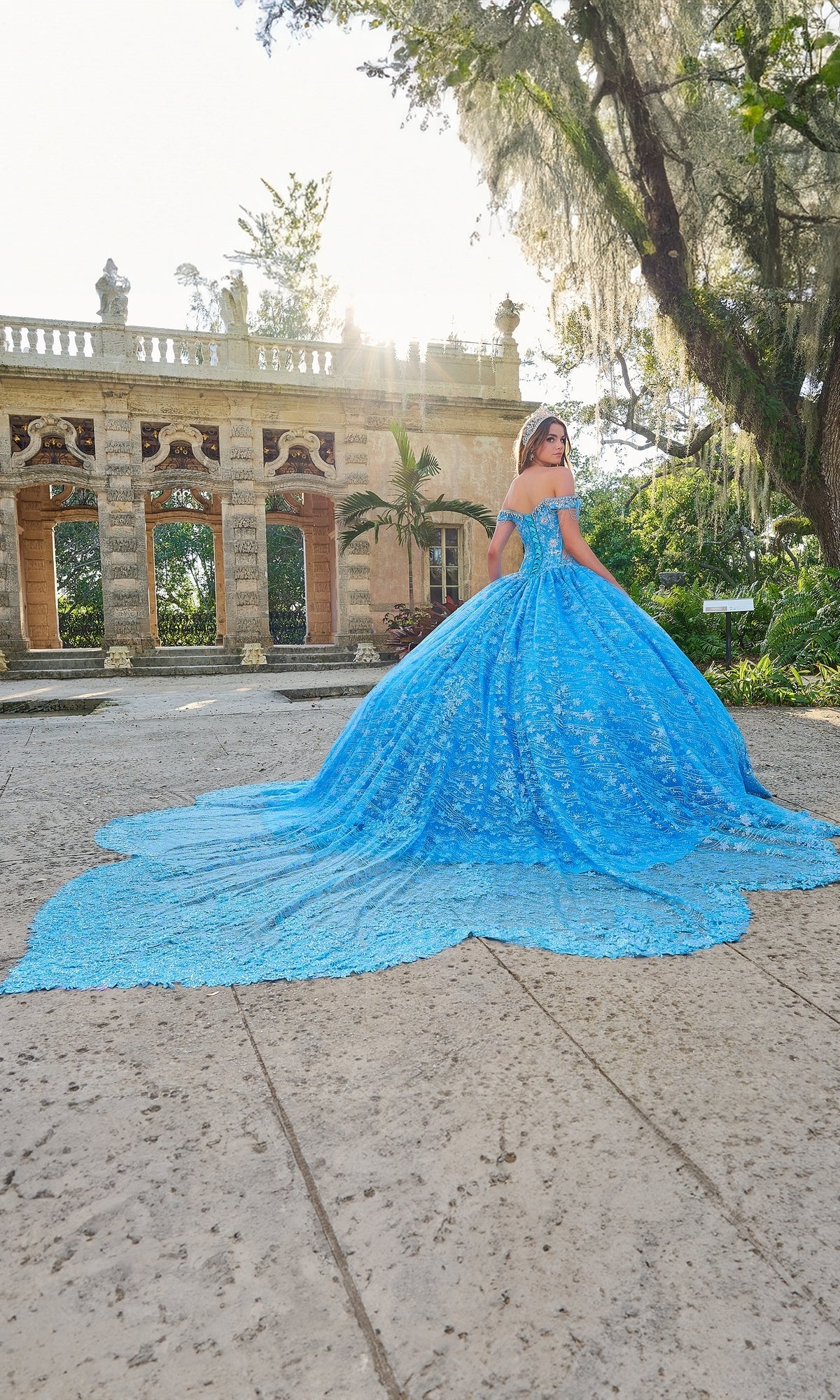A woman in the Quinceanera Dress 54220 by Amarra, featuring an off-the-shoulder neckline and long train, stands outdoors before a historic stone building and trees.