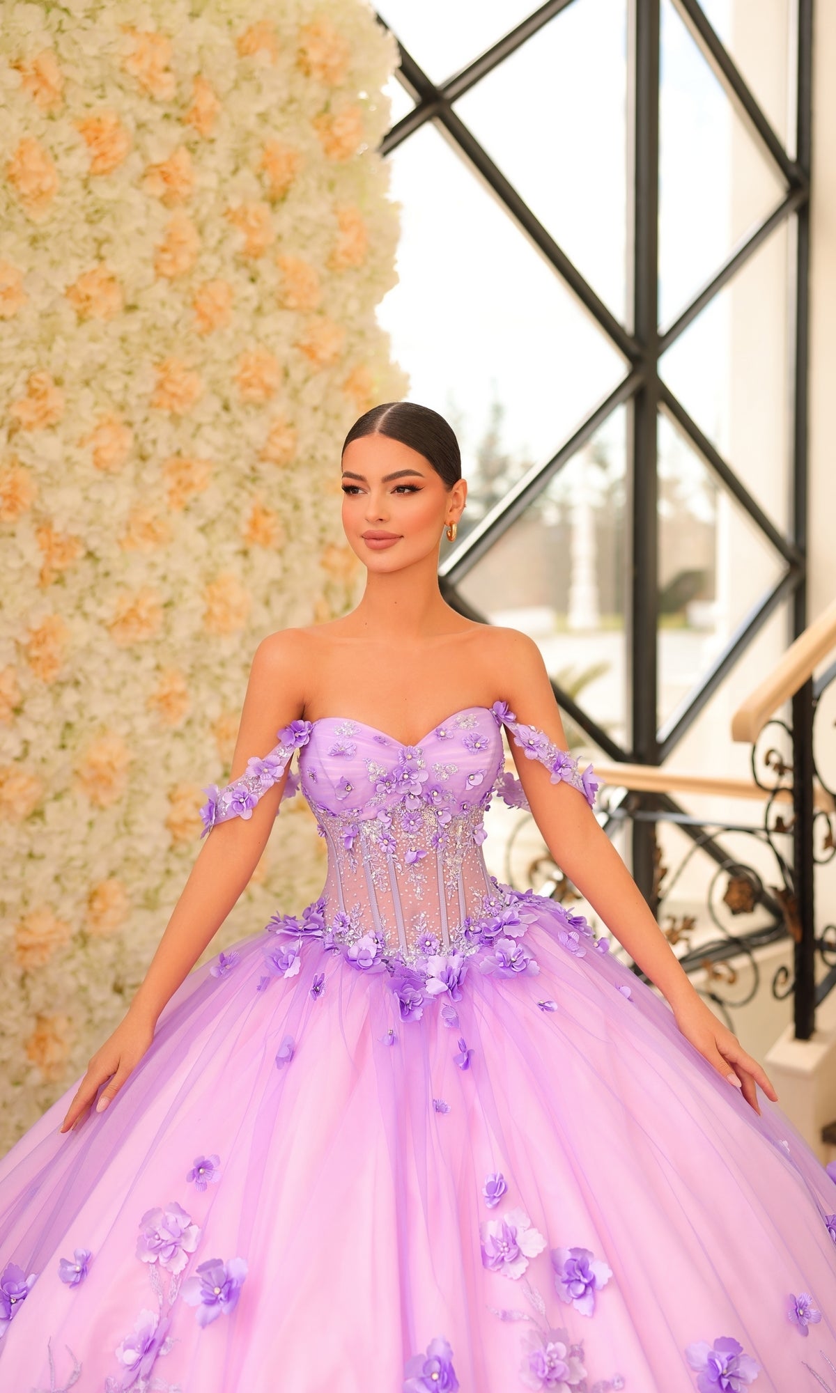 A woman wearing the Quinceanera Dress 54224 By Amarra, an off-the-shoulder lavender tulle gown with floral appliqué, stands indoors by a wall of white and peach flowers, her dress beautifully complementing the scene.