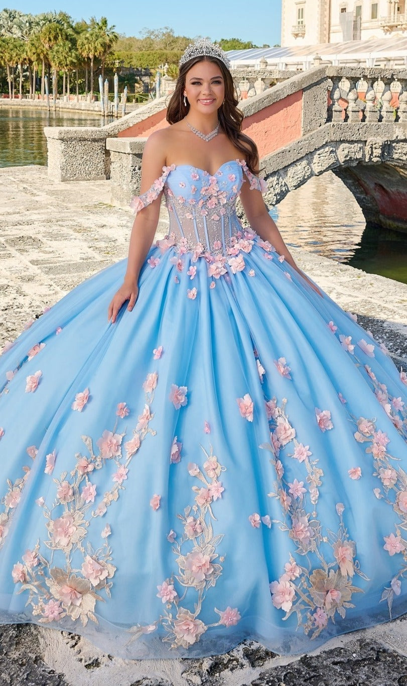 A young woman stands outdoors by a stone staircase, wearing the Quinceanera Dress 54224 By Amarra— a blue tulle ball gown with pink floral appliqué—paired with a tiara and necklace.