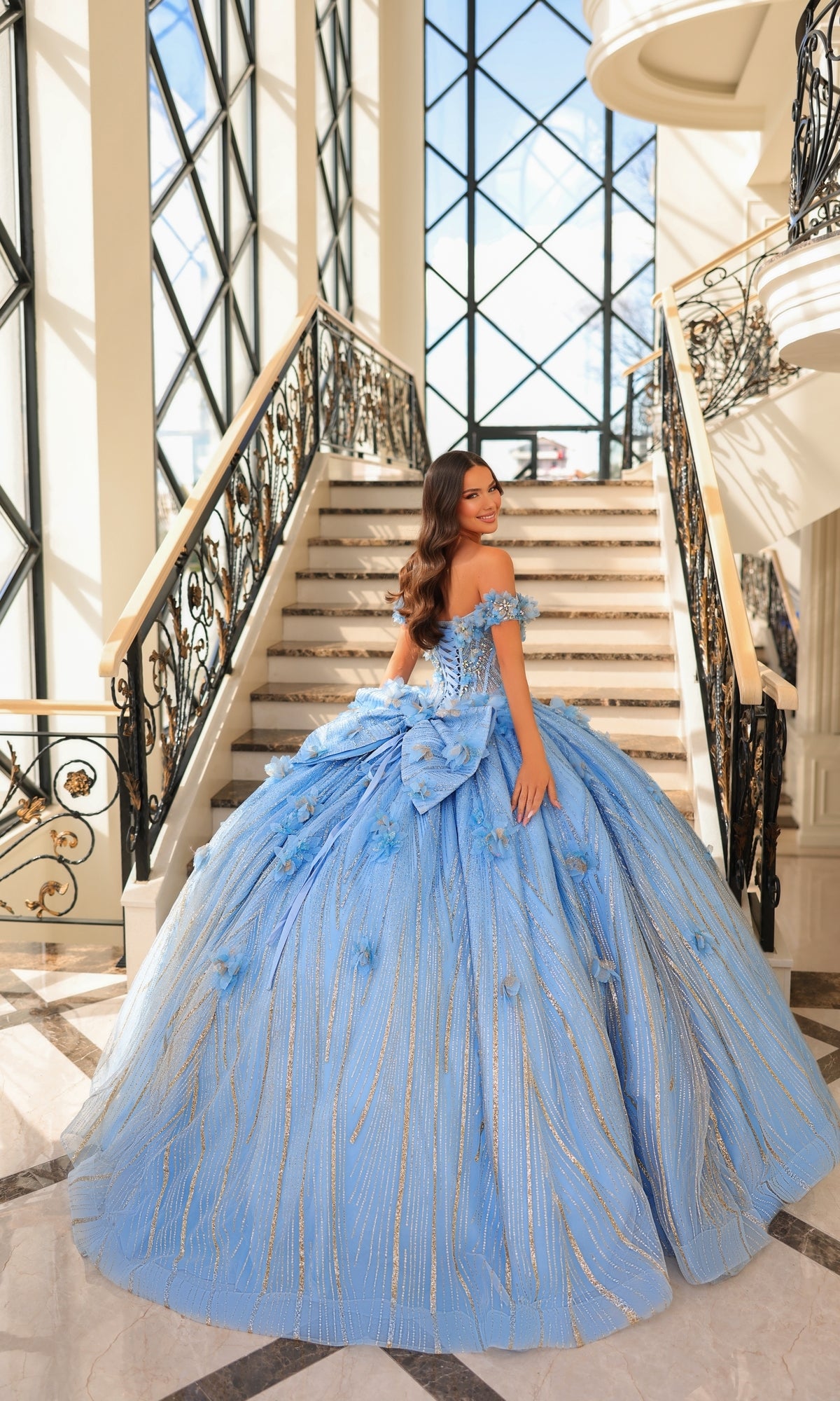 A woman in the Quinceanera Dress 54226 By Amarra, a voluminous light blue ball gown with floral appliqué, stands on an elegant staircase with decorative railings in a sunlit, grand interior.