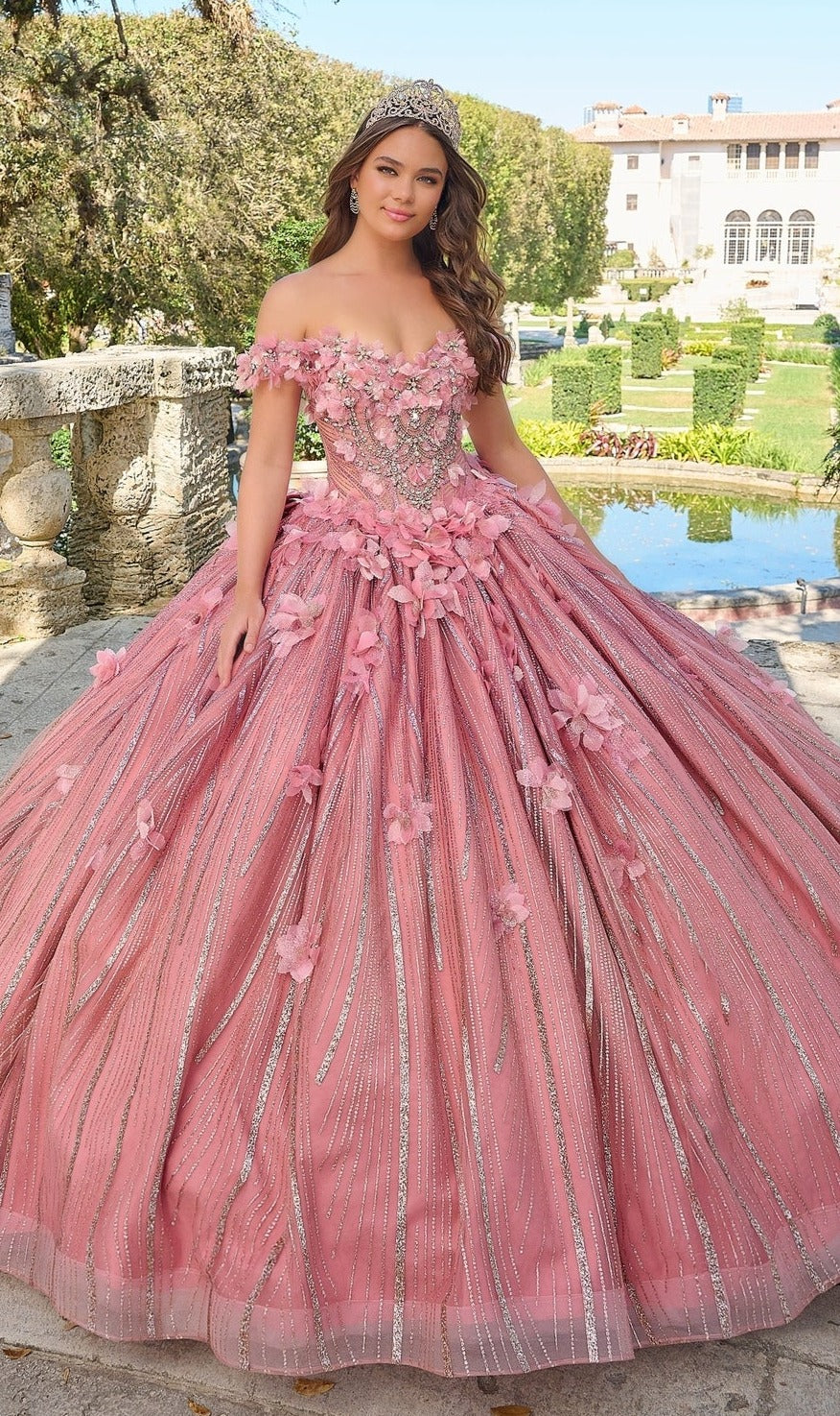 A young woman in the Quinceanera Dress 54226 by Amarra, featuring ornate floral appliqué and an off-the-shoulder design, stands outdoors in a garden with stone structures and a large building behind her.
