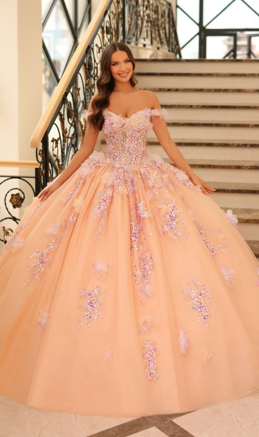 A young woman smiles on a staircase, wearing the Quinceanera Dress 54228 by Amarra—a peach ball gown with 3D floral appliqué and off-the-shoulder sleeves.