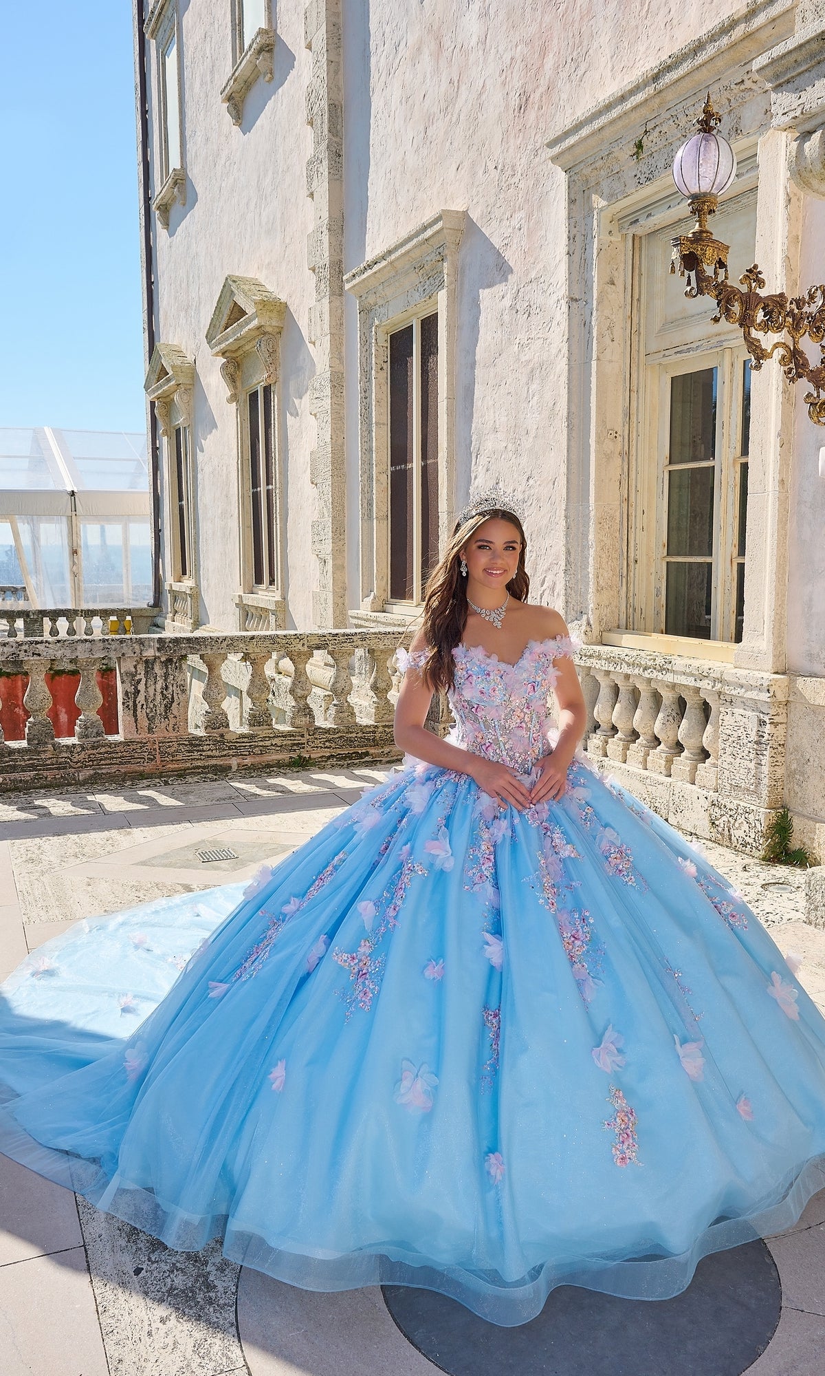 A young woman wears the Quinceanera Dress 54228 by Amarra, an off-the-shoulder blue ball gown with 3D floral appliqué, as she stands on an outdoor stone balcony near an ornate building in daylight.