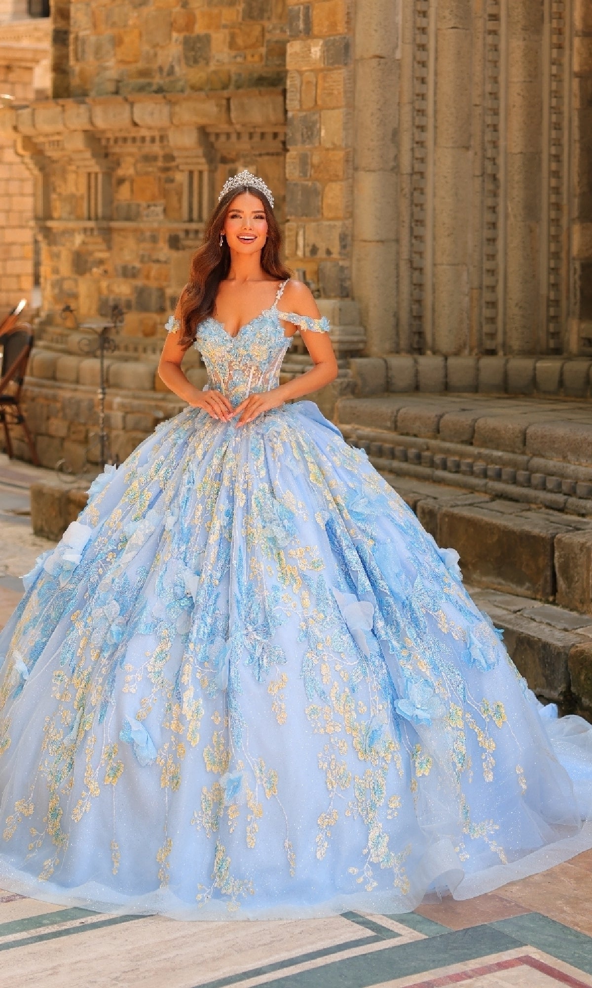 A woman in the Sheer-Corset Quinceanera Dress: Amarra 54232, adorned with 3D floral appliques and a tiara, stands before a stone building with ornate architecture.