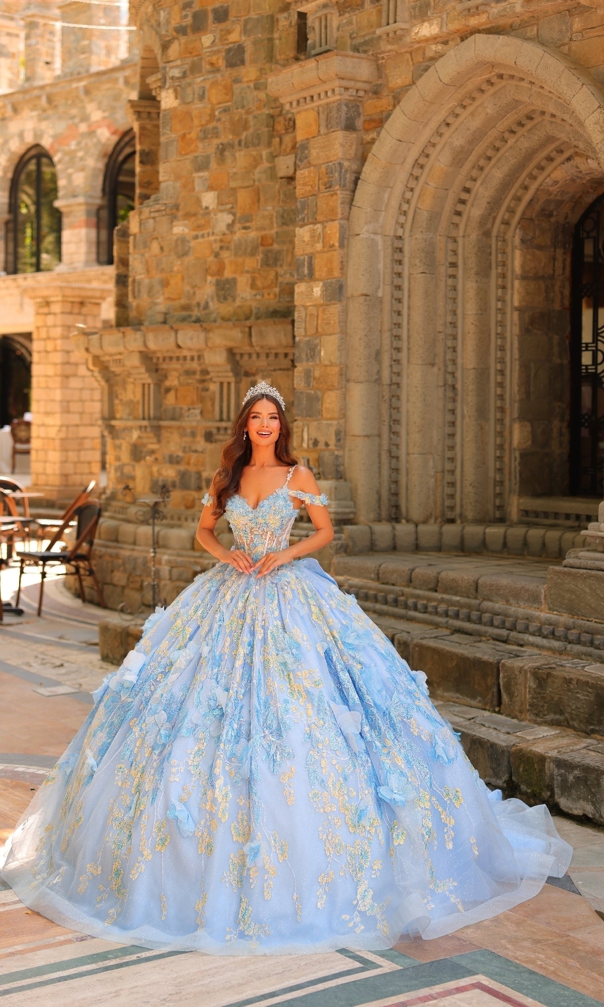 A woman in the Sheer-Corset Quinceanera Dress: Amarra 54232, featuring pastel hues and 3D floral appliques with a tiara, stands before a historic stone building with arched doorways.