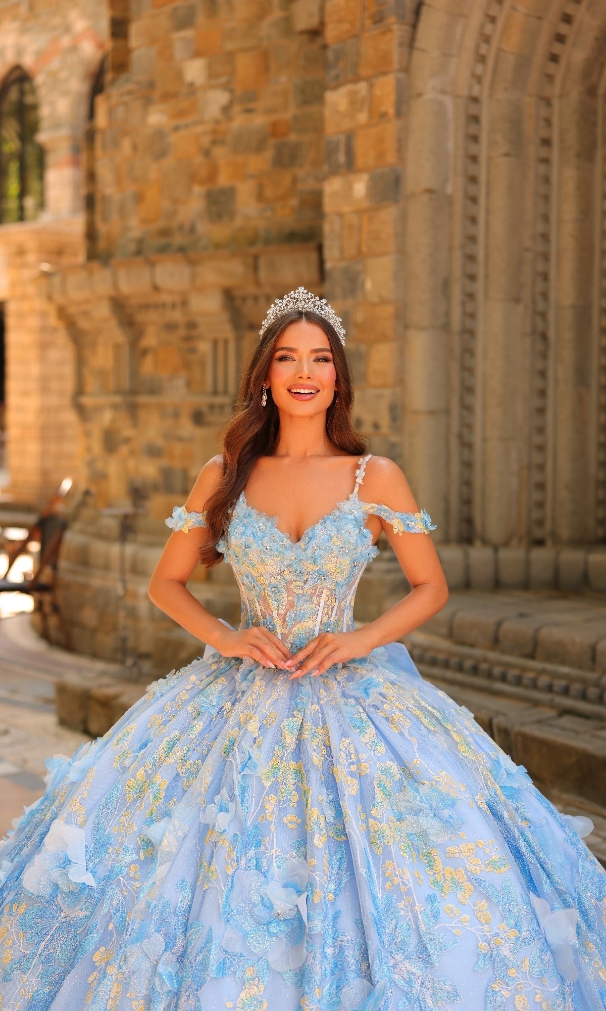 A woman in the Sheer-Corset Quinceanera Dress: Amarra 54232 with 3D floral appliques and a tiara stands smiling, hands clasped in front of her waist, in front of a stone building.