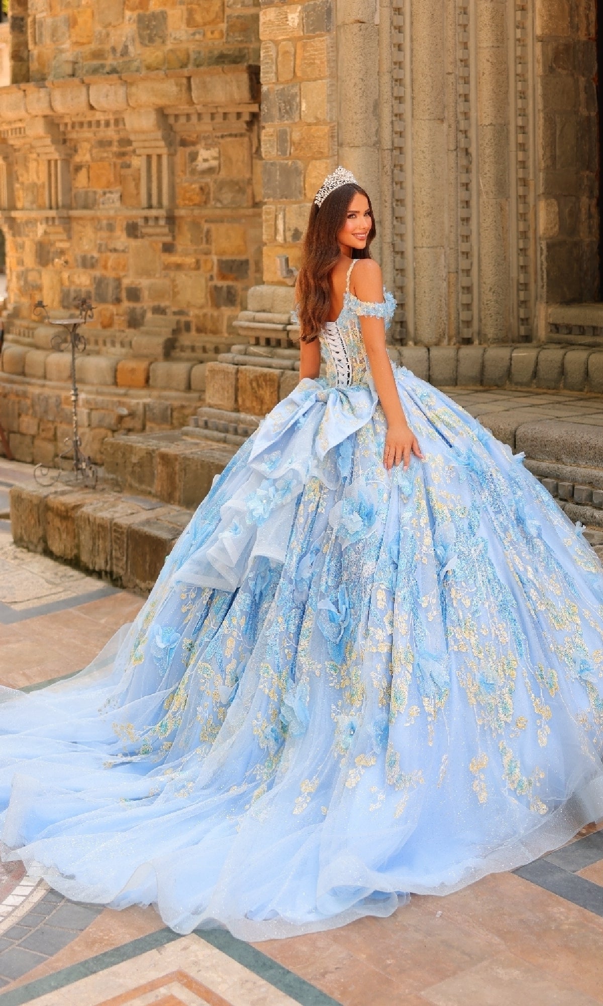 A woman in the Sheer-Corset Quinceanera Dress: Amarra 54232 with 3D floral appliques and a tiara stands before an ornate stone building, glancing over her shoulder.