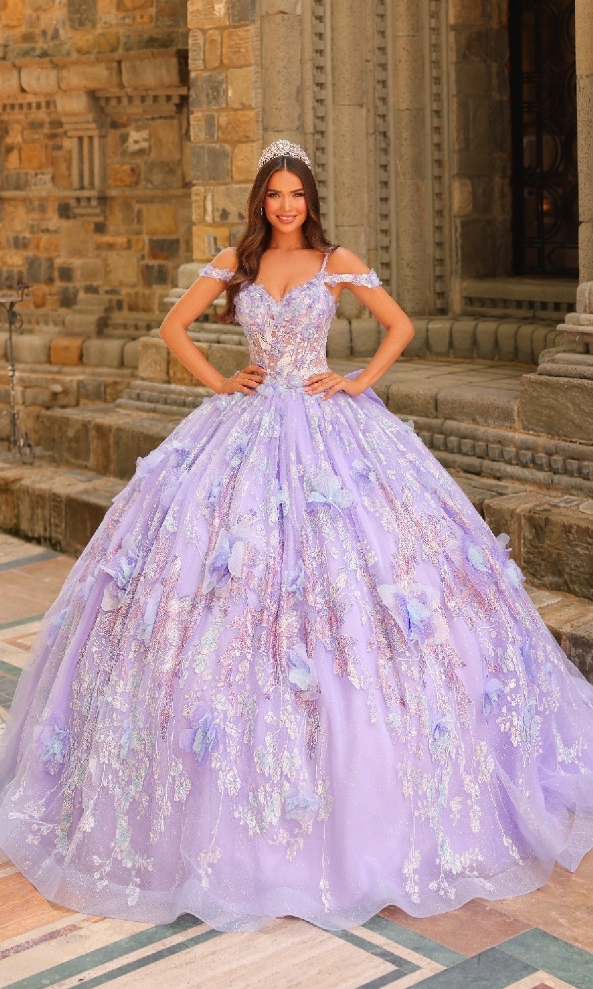 A woman in the Amarra 54232 Sheer-Corset Quinceanera Dress, a sparkling lavender ball gown with a sheer corset bodice, stands before an ornate stone building wearing a tiara.