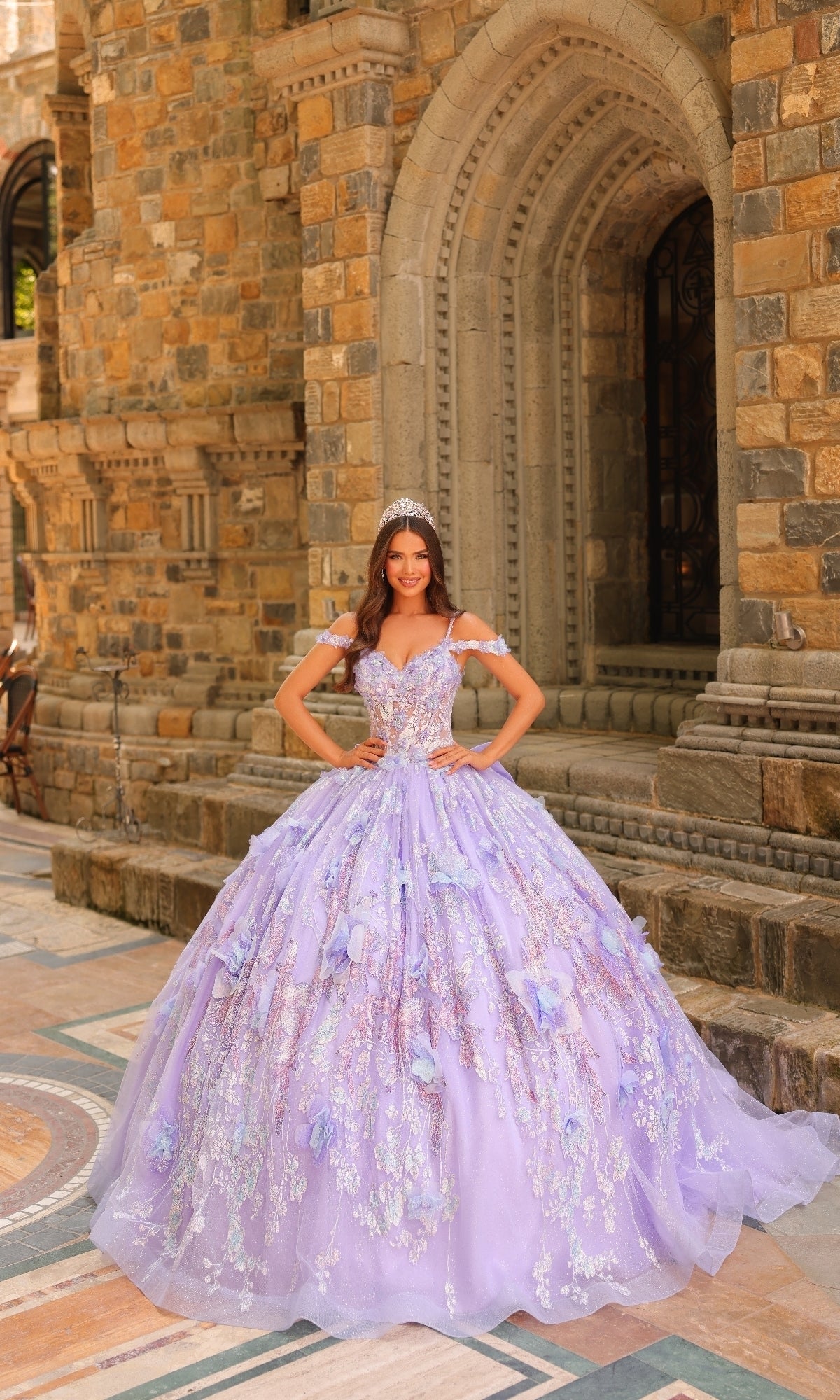 Wearing the Sheer-Corset Quinceanera Dress: Amarra 54232, a woman in a lilac gown with 3D floral appliques and a tiara stands before a stone building with arched doorways.