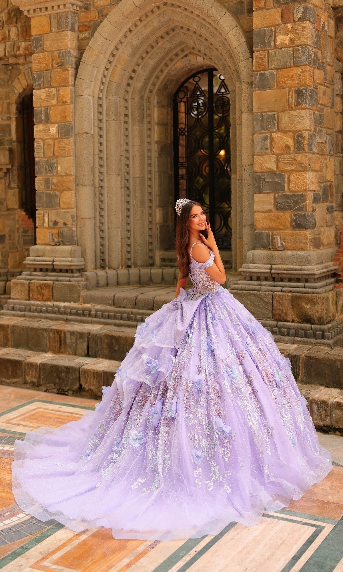 A woman in the Sheer-Corset Quinceanera Dress: Amarra 54232 with a tiara stands before a stone building with arched doorways, glancing over her shoulder.