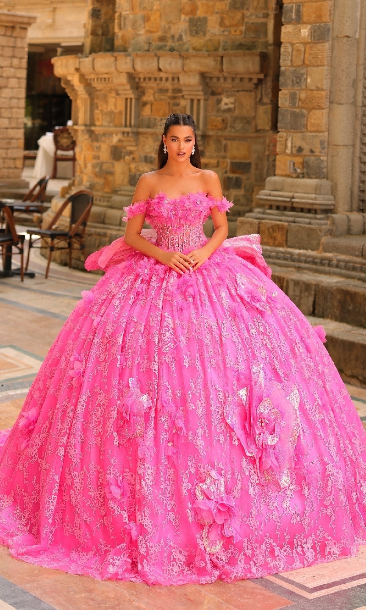 A young woman stands outdoors before stone architecture, wearing the Amarra 54233 3D Flower Quinceanera Dress with off-the-shoulder sleeves, detachable straps, lace details, and bold pink 3D flower decorations.
