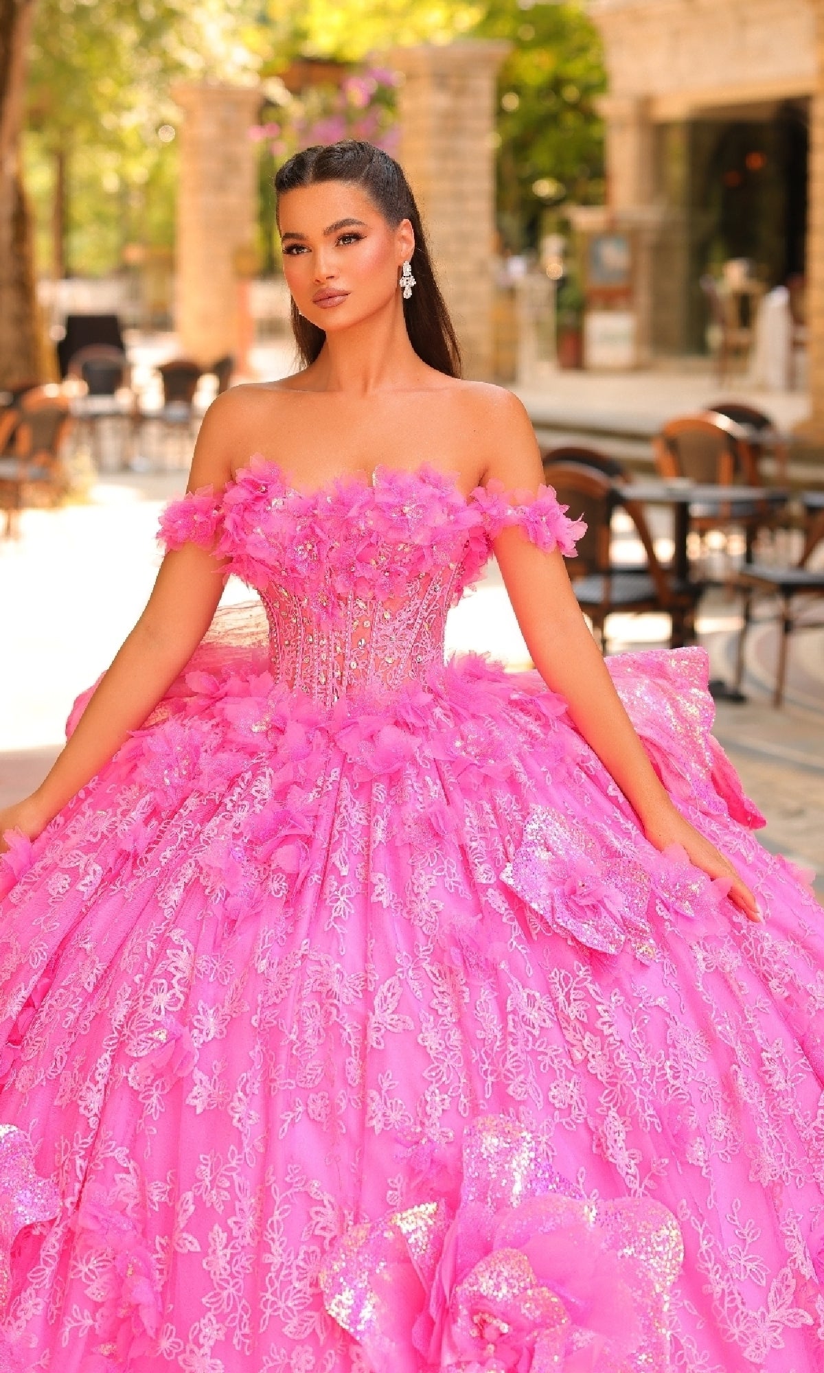 A woman poses outdoors near tables and chairs, wearing the Amarra 54233—a bright pink quinceanera ball gown featuring 3D flowers and detachable straps.