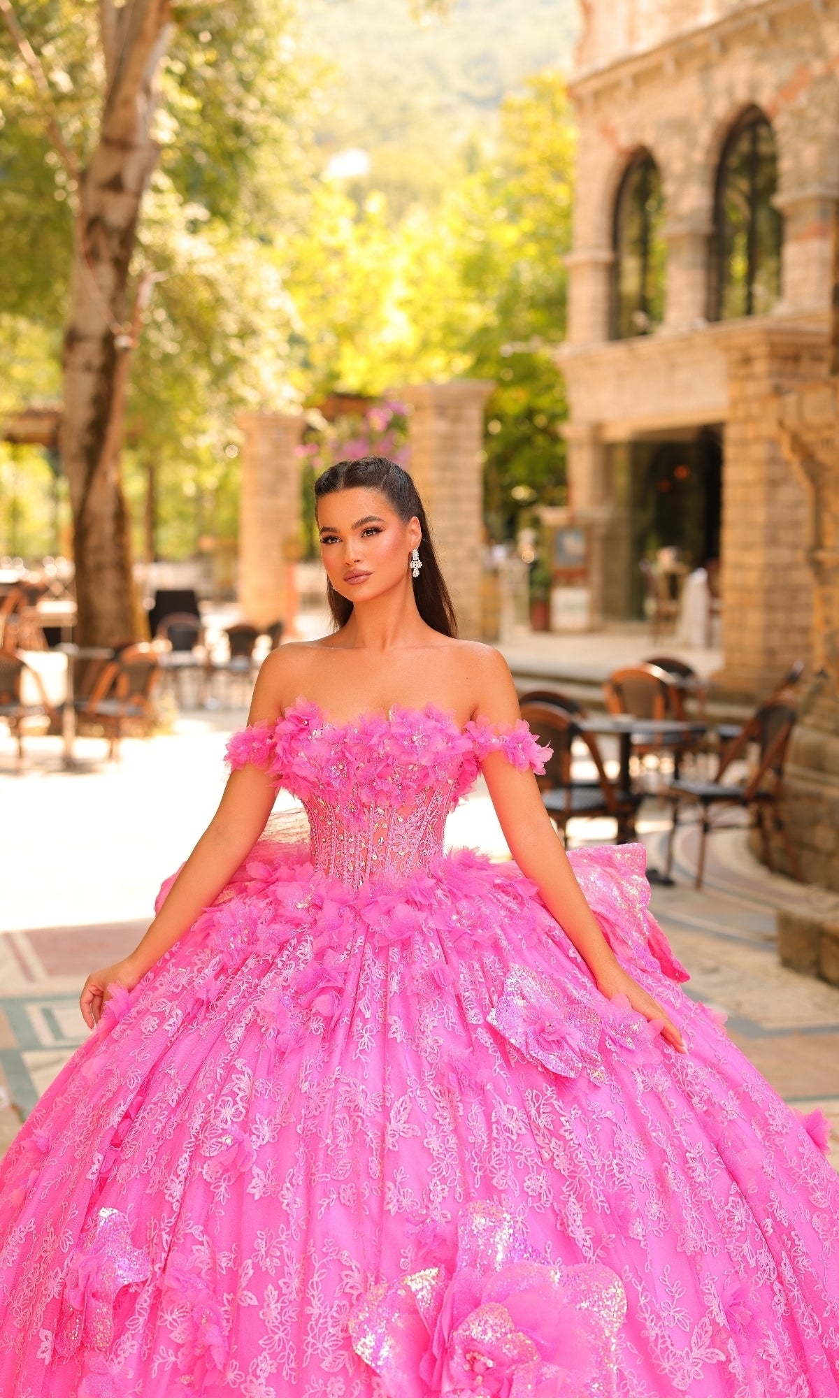 A woman stands outdoors in the Amarra 54233 3D Flower Quinceanera Dress, featuring a voluminous off-the-shoulder ball gown with an embellished bodice. Trees and stone buildings are visible in the background.