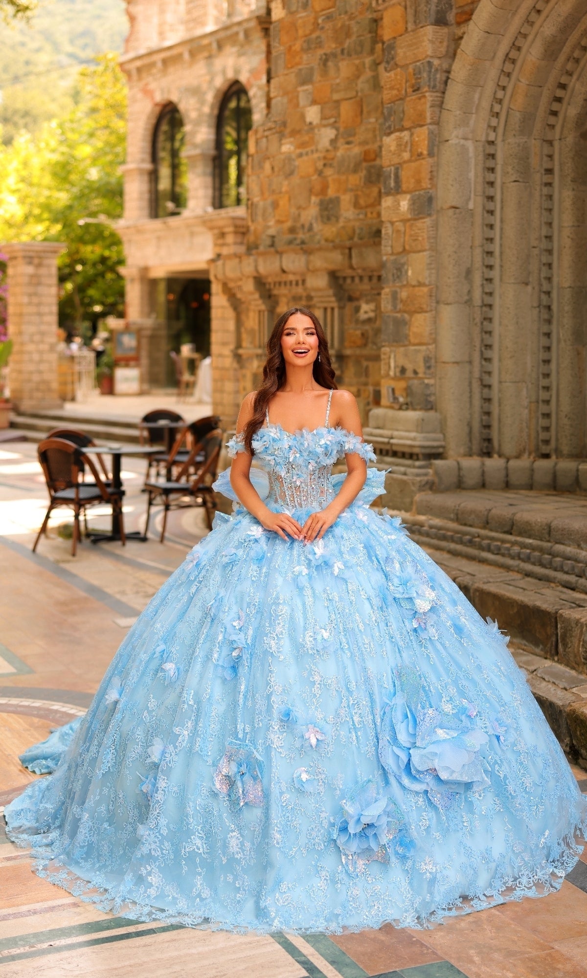 Wearing the Amarra 54233 3D Flower Quinceanera Dress with detachable straps, a woman smiles outdoors in front of a stone building, showcasing the gown's elaborate blue design and delicate floral accents.
