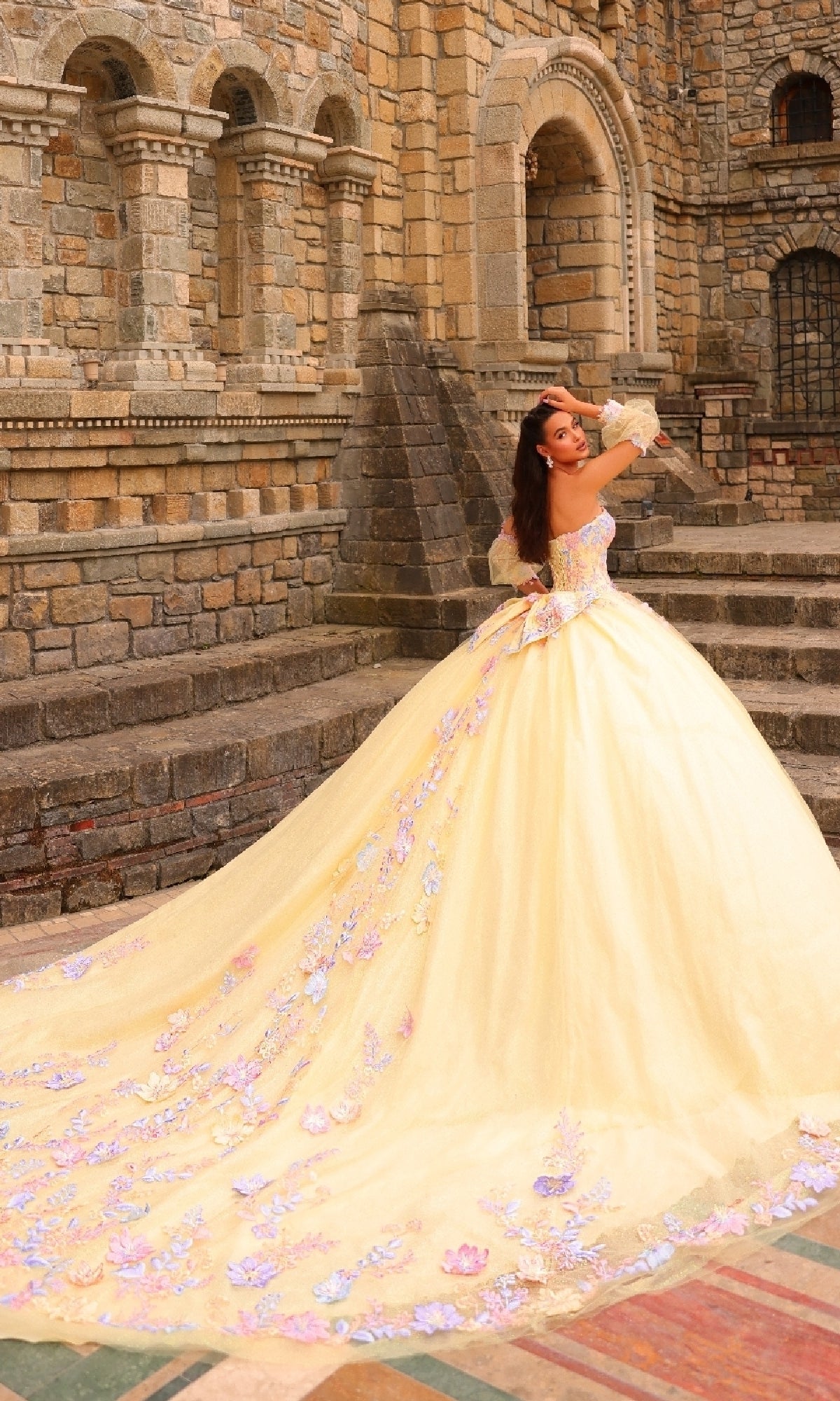 Wearing the Amarra 54234 Puff-Sleeve Quinceanera Dress in pale yellow with a long train and floral lace appliqués, a woman stands on stone steps outside a historic building.