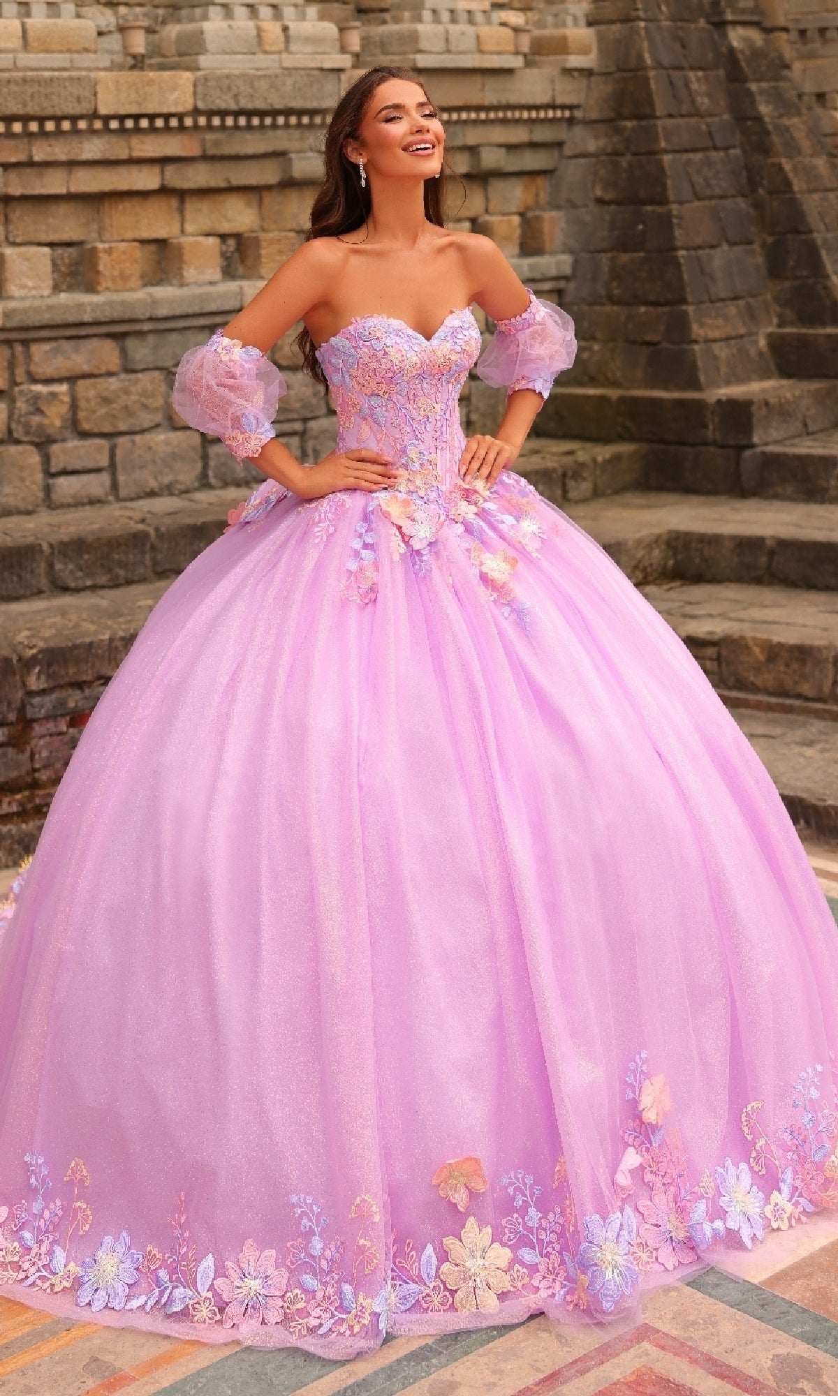 A woman in the Amarra 54234 Puff-Sleeve Quinceanera Dress, a light purple gown with floral lace appliqués, stands outdoors before stone steps and a brick wall.