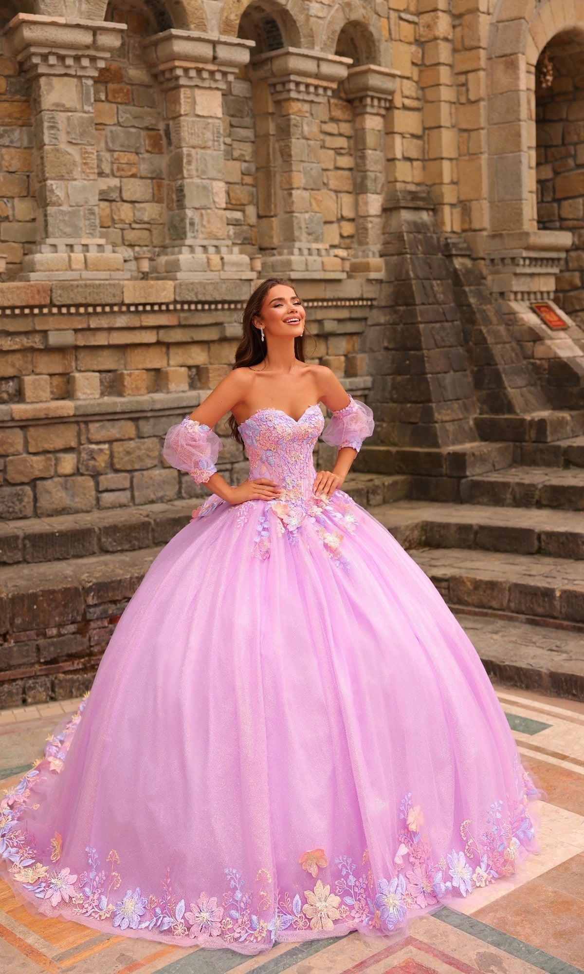 A woman smiles in front of a stone building, wearing the Amarra 54234 Puff-Sleeve Quinceanera Dress in pink, featuring delicate floral lace appliqués.
