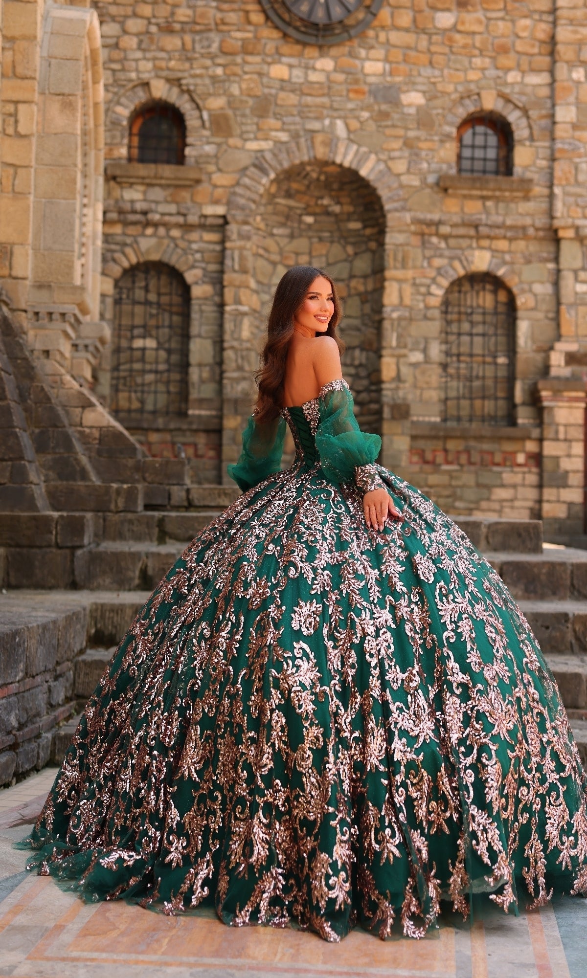 A woman stands outdoors in front of a stone building, wearing the Sequin Quinceanera Dress: Amarra 54238, an off-shoulder green ball gown with gold embroidery, a corset bodice, and a voluminous skirt.