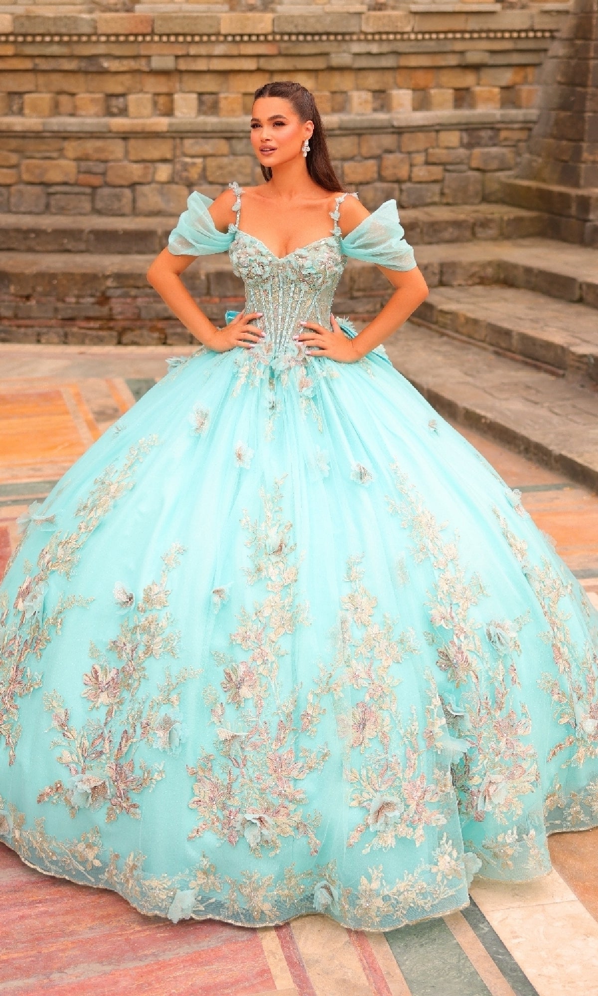 A woman wearing the Puff-Sleeve Quinceanera Dress: Amarra 54241, featuring light blue fabric, 3D flowers, and floral embroidery, stands on a patterned floor with stone steps and a brick wall behind her.