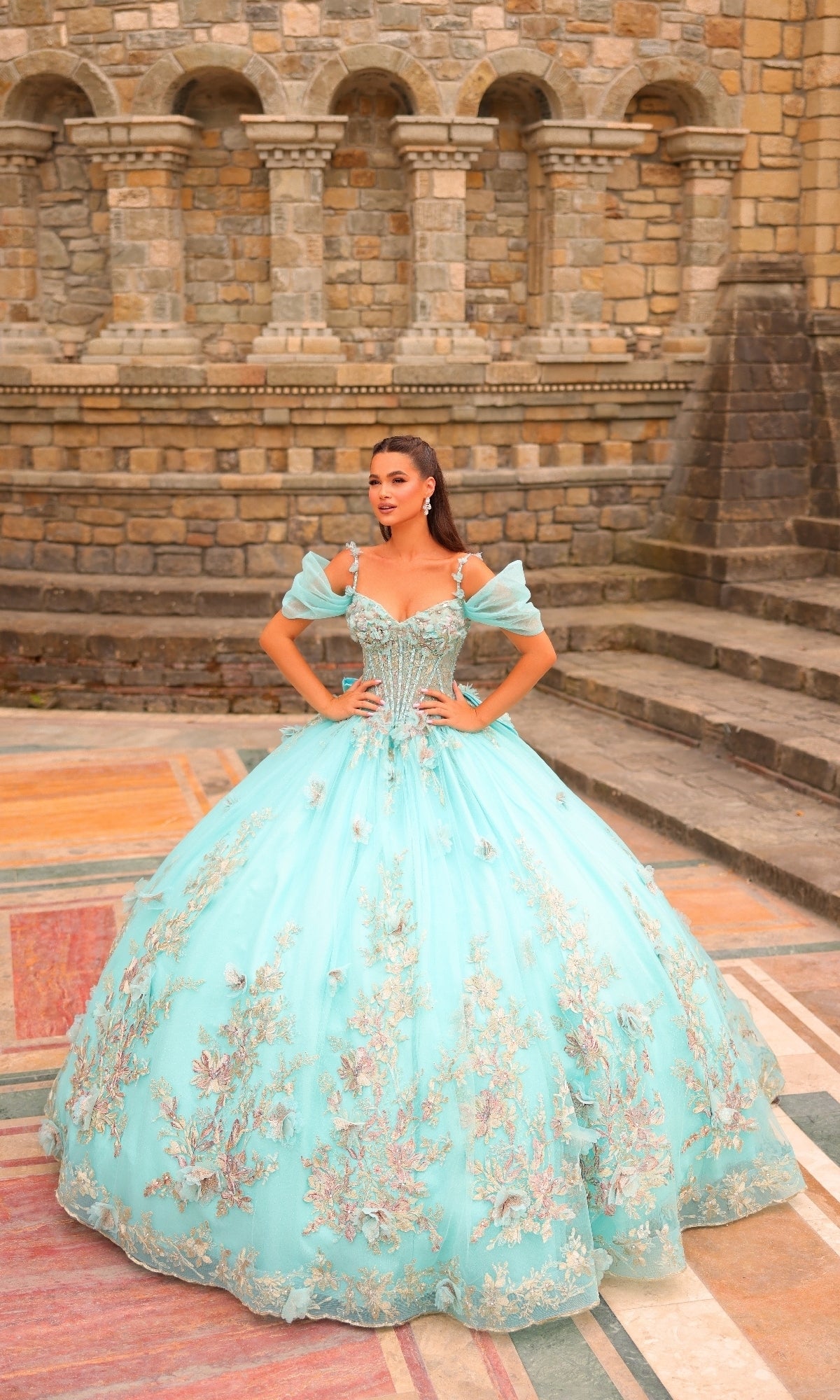 A woman wearing the Amarra 54241 Puff-Sleeve Quinceanera Dress with glitter tulle and 3D flowers stands on a colorful stone courtyard in front of a castle-like wall.