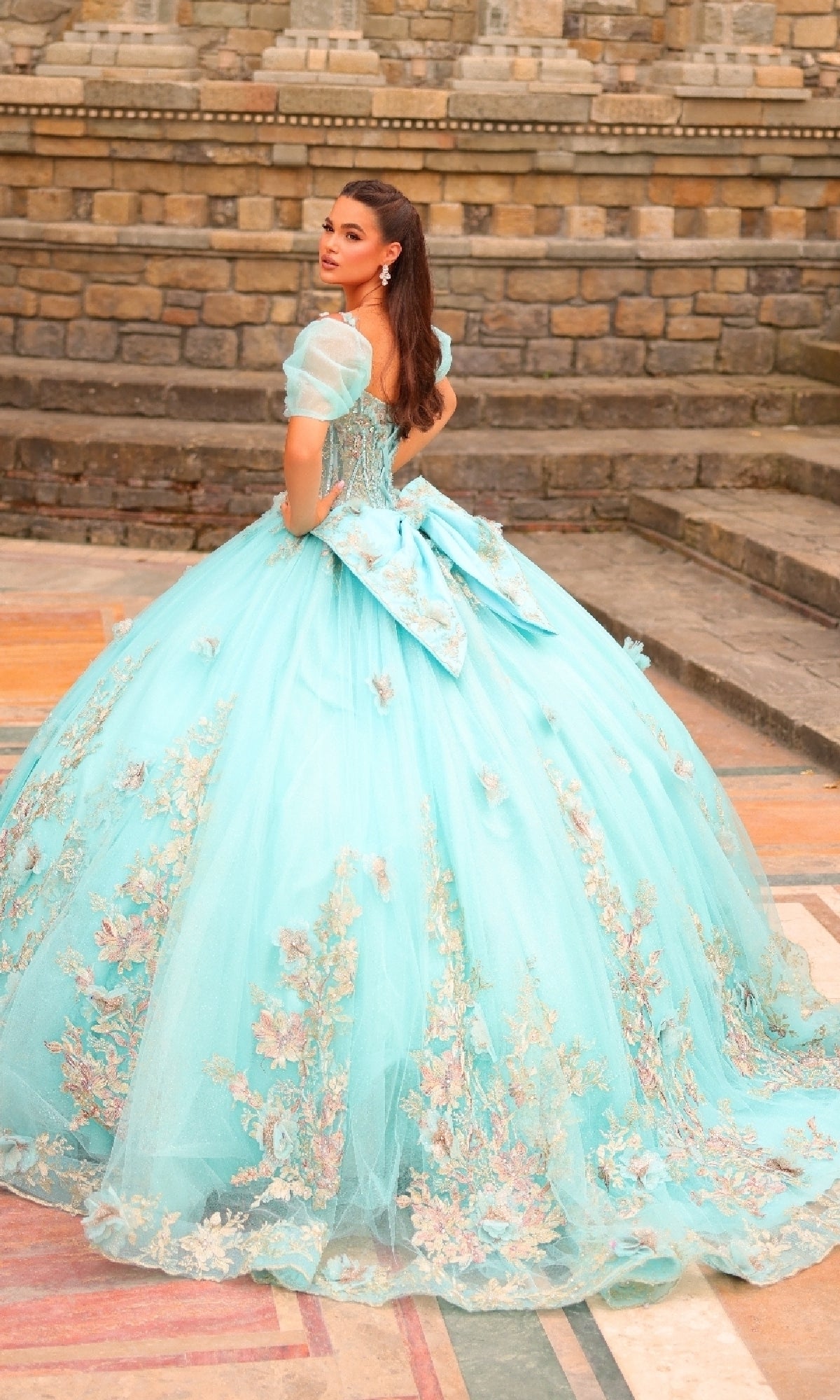 A woman in the Amarra 54241 Puff-Sleeve Quinceanera Dress, featuring light blue lace and 3D flowers, stands on patterned stone steps and looks over her shoulder.