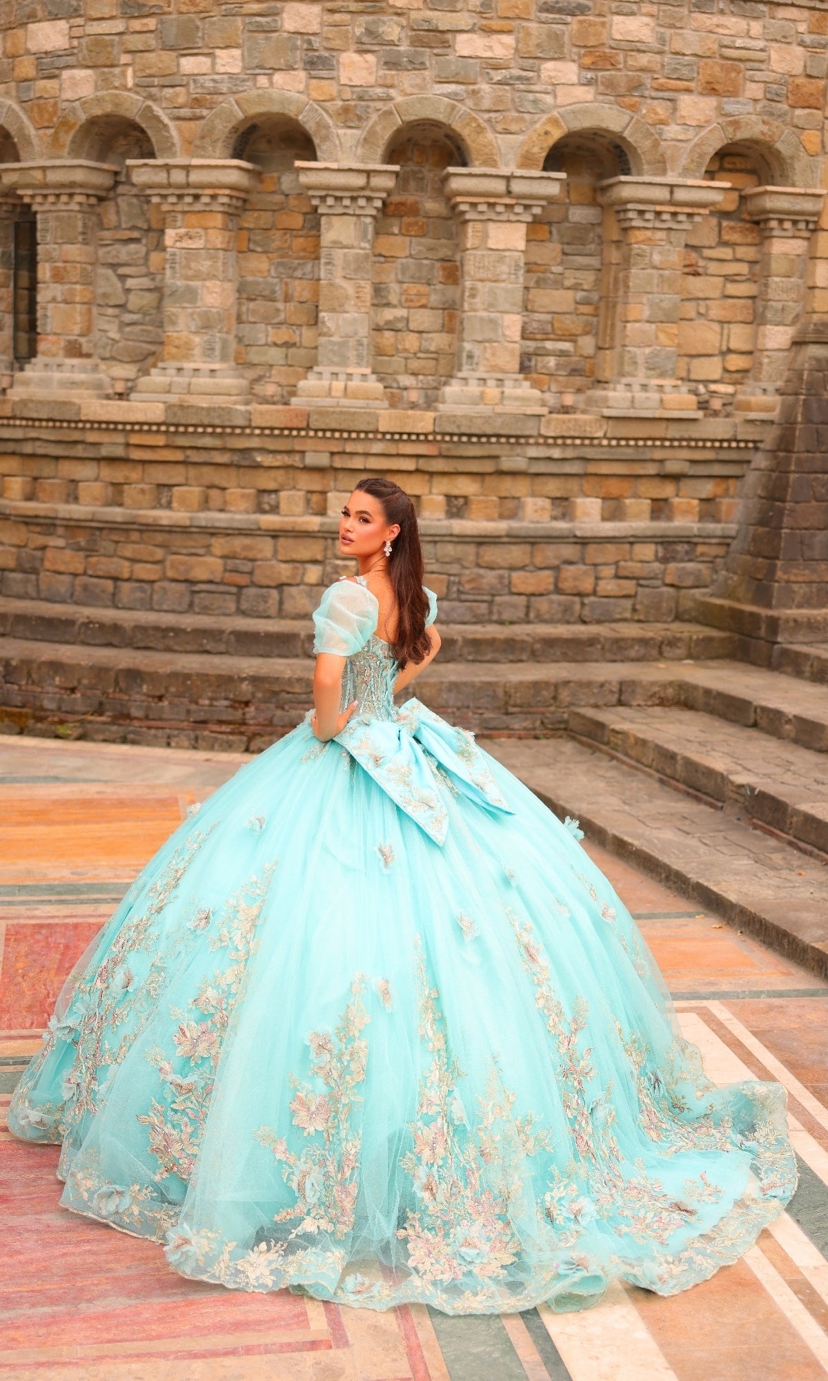 A woman in the Amarra 54241 Puff-Sleeve Quinceanera Dress with 3D blue floral details stands outdoors before a stone wall, glancing back over her shoulder.