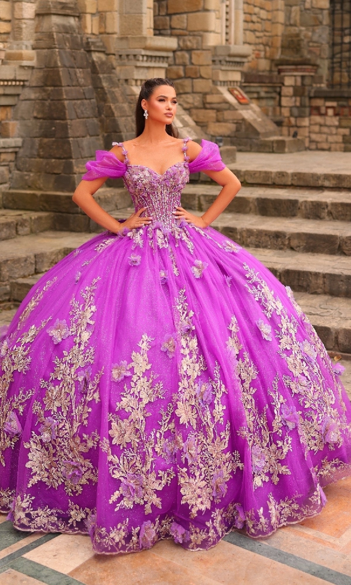 A woman in the Amarra 54241 Puff-Sleeve Quinceanera Dress, featuring bright purple fabric, gold floral embroidery, and glitter tulle, stands on stone steps outside a historic building.