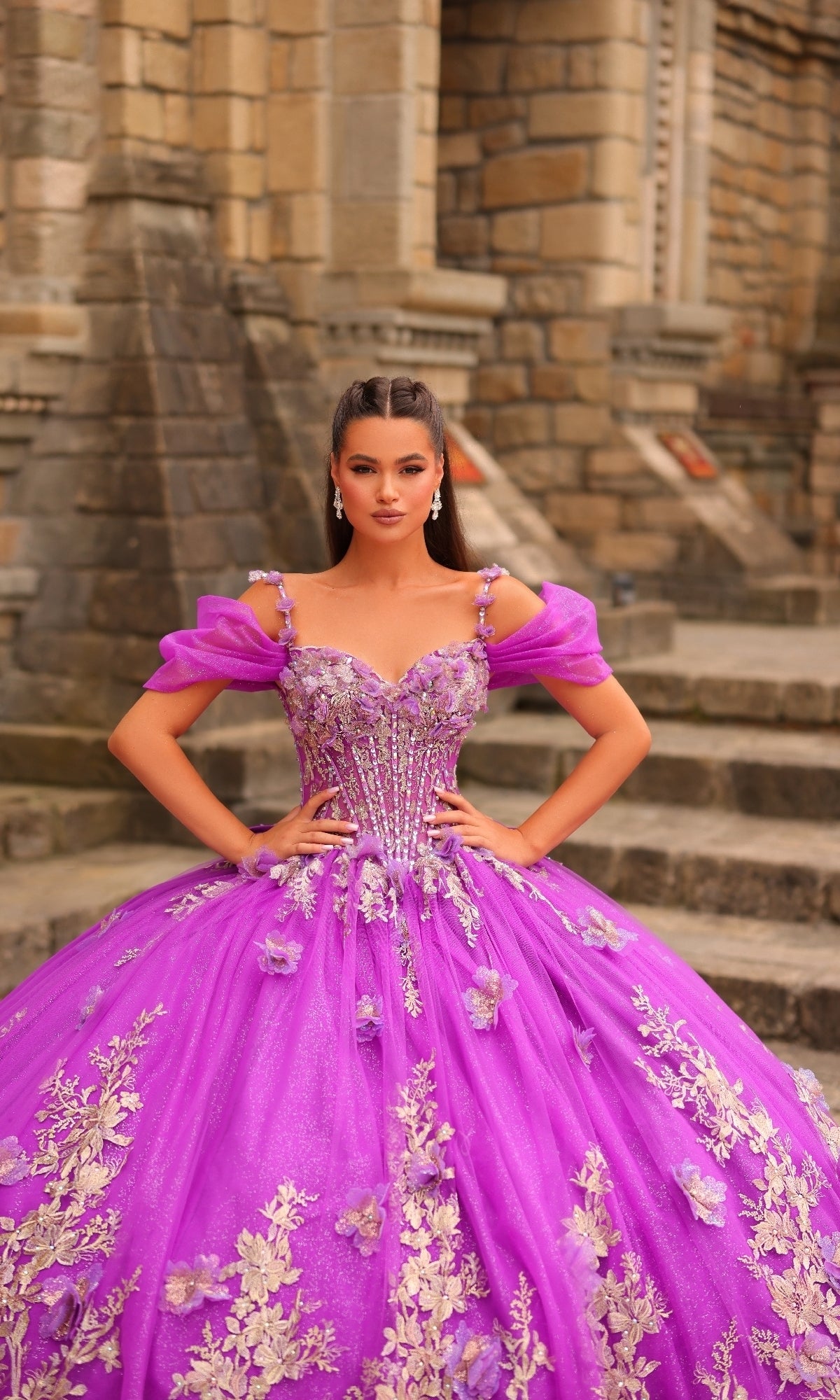 A woman in the Amarra 54241 puff-sleeve quinceanera dress, featuring bright purple fabric with gold floral detailing and 3D flowers, stands before a stone building with steps and columns.