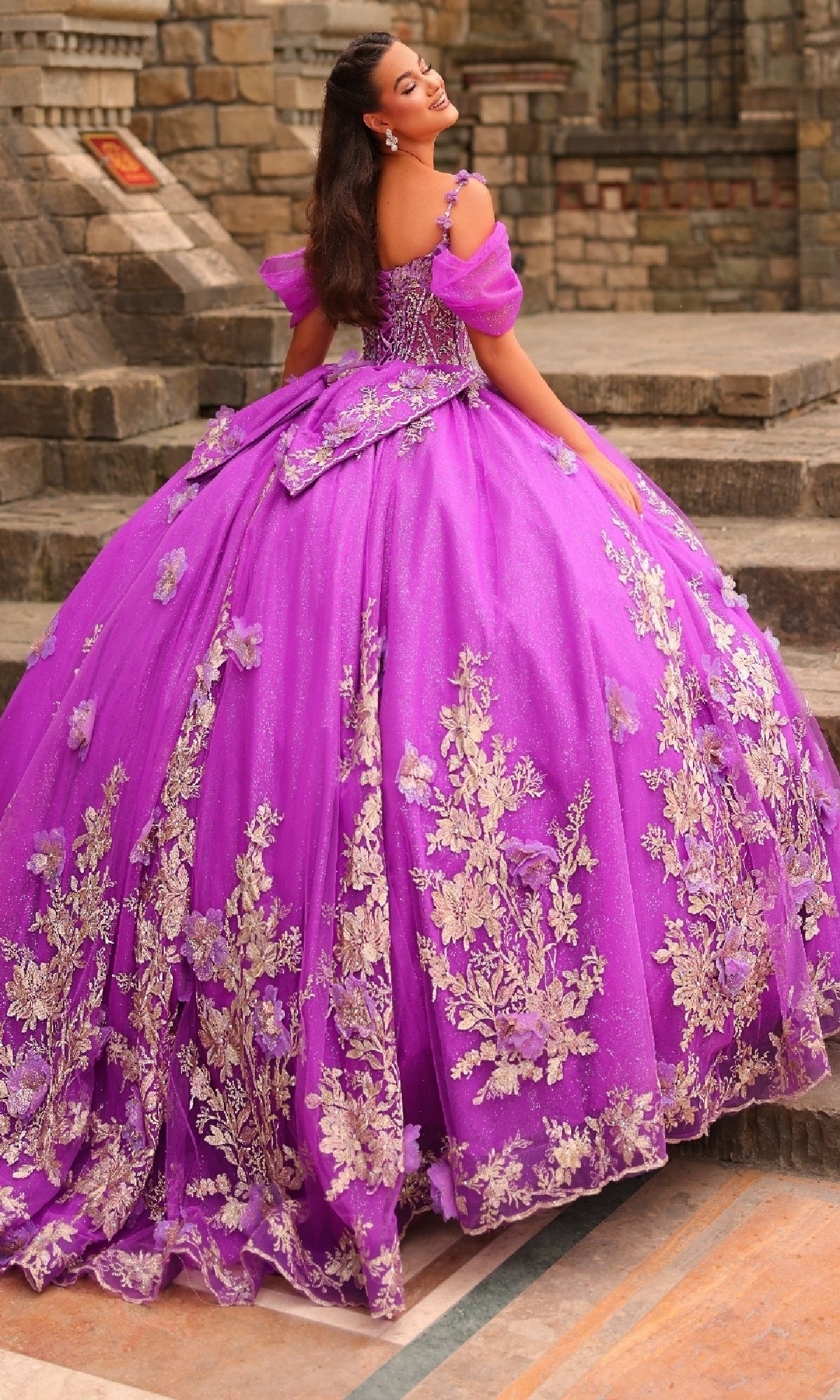 A woman stands on stone steps outdoors wearing the Puff-Sleeve Quinceanera Dress: Amarra 54241, featuring vibrant purple fabric, gold floral embroidery, glitter tulle accents, puff sleeves, and a large back bow.