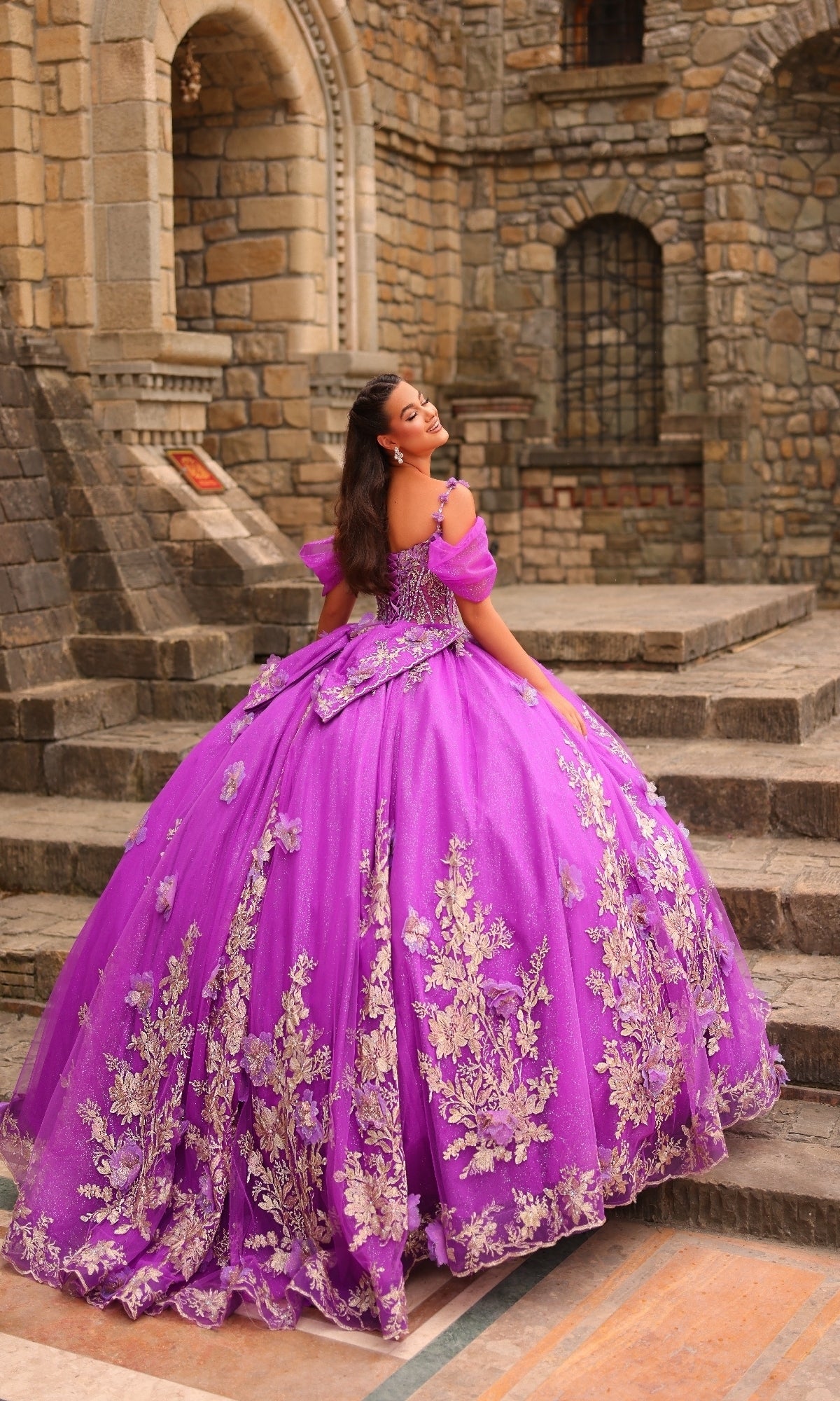 A woman in the Puff-Sleeve Quinceanera Dress: Amarra 54241, featuring bright purple fabric, gold floral embroidery, and glitter tulle, stands on stone steps before an old building, looking over her shoulder and smiling.