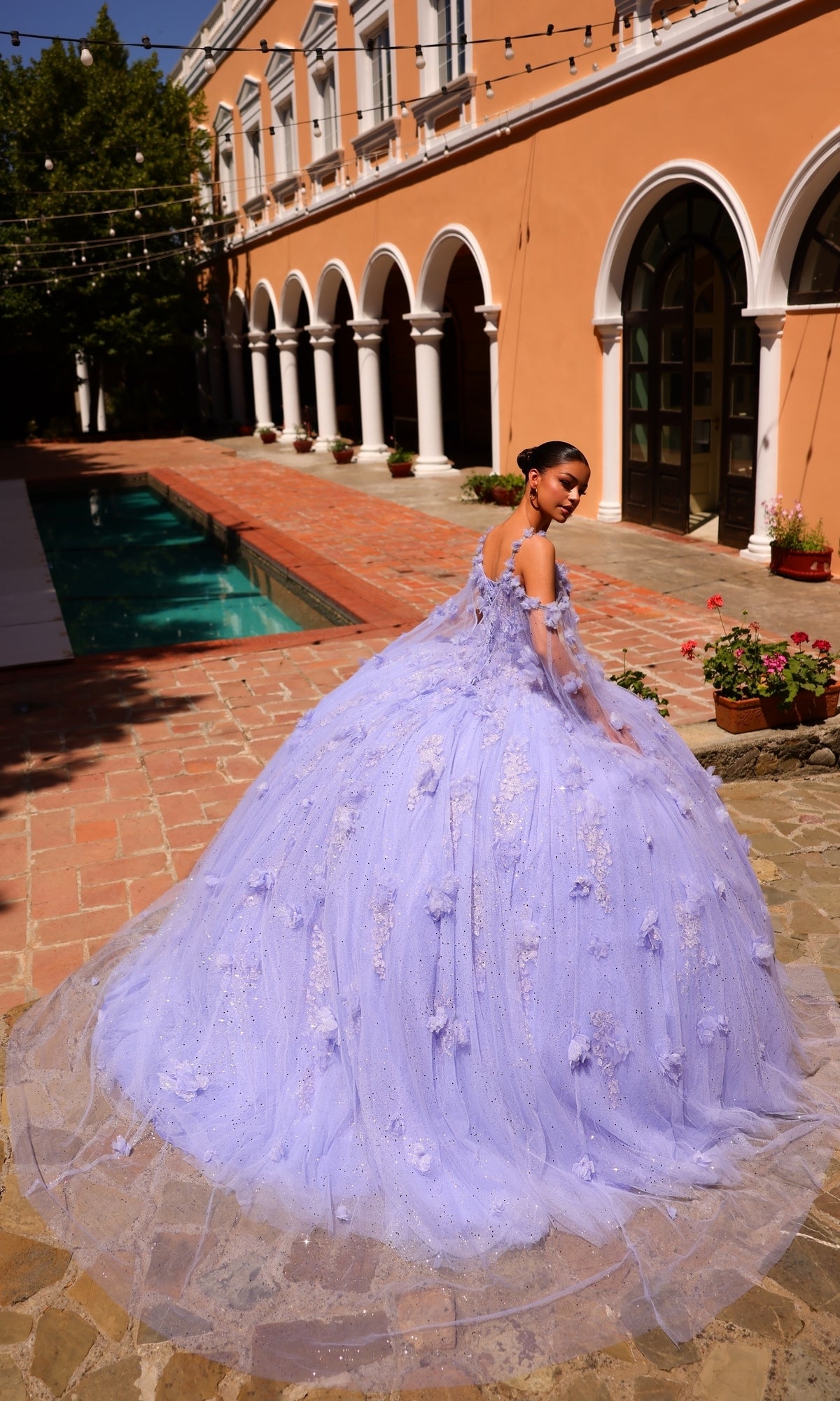 A young woman wears the Amarra 54254 Periwinkle Purple Quincea¤era Dress, featuring voluminous embroidery and glitter, as she stands by a brick walkway and pool outside a peach-colored building with arches.