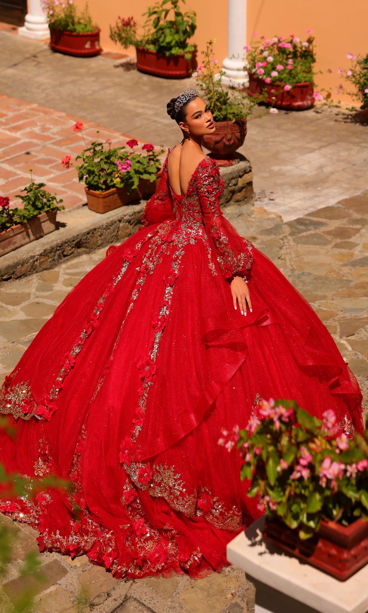 A woman in the Amarra 54263 Red Quincea¤era Dress with bell sleeves and a sparkling tiara stands on an outdoor stone path, surrounded by potted flowering plants.