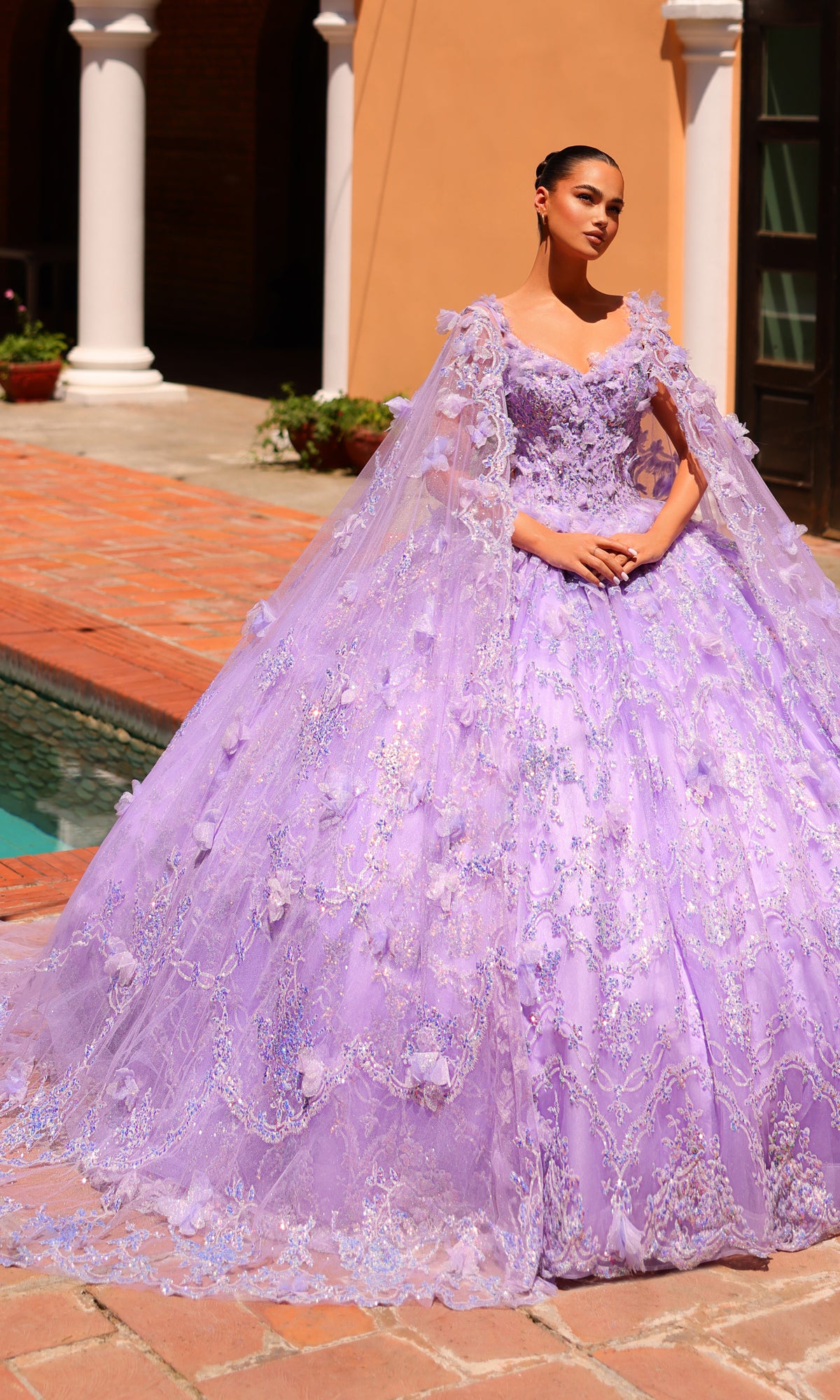 A woman stands by a pool outside an arch-adorned building, wearing the Amarra 54281 Lilac Purple Quincea¤era Dress with floral embellishments and glitter tulle.