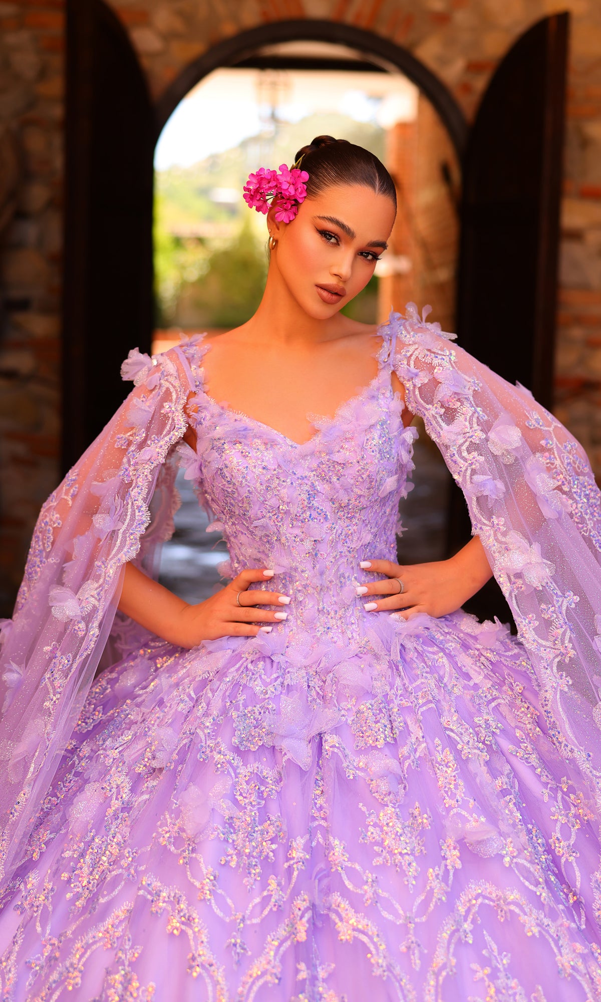 A woman poses in front of a stone archway wearing the Amarra 54281 Lilac Purple Quincea¤era Dress, featuring floral accents and matching cape sleeves, with pink flowers adorning her hair.