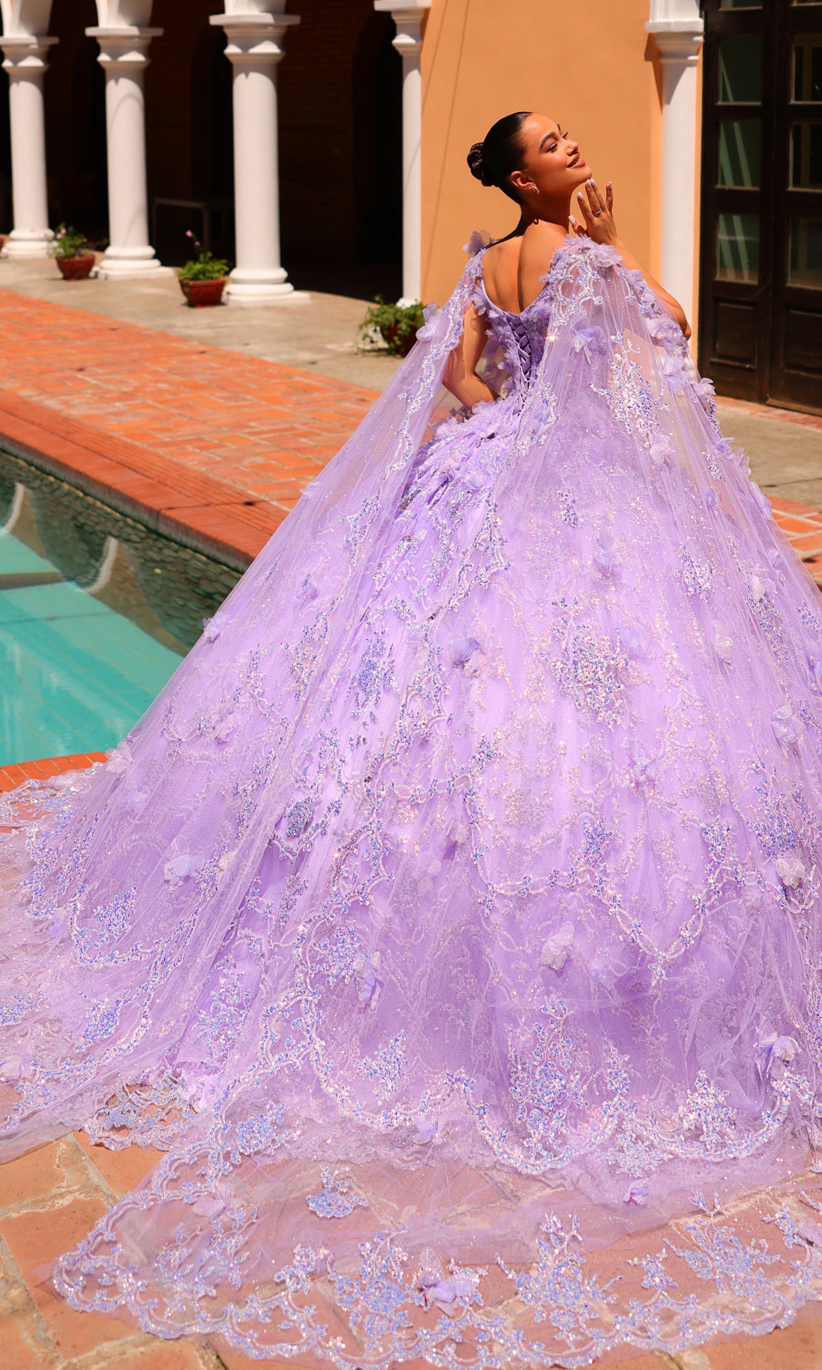 A woman poses by a pool in the Amarra 54281 Lilac Purple Quincea¤era Dress, featuring a voluminous silhouette, long ornate lace train, and cape, with one hand near her face.