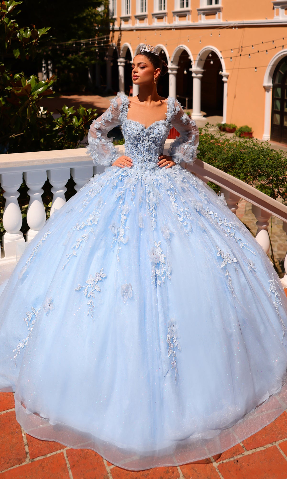 A woman wearing the Corset Ball Gown Quinceañera Dress 59290 by Amarra and a tiara stands on a balcony with Mediterranean-style architecture in the background.