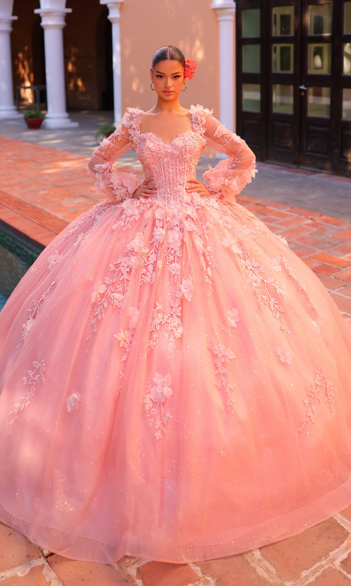 A young woman poses outdoors near a tiled courtyard and white columns in the Corset Ball Gown Quinceañera Dress 59290 by Amarra, featuring glitter tulle, long sleeves, and a full skirt.