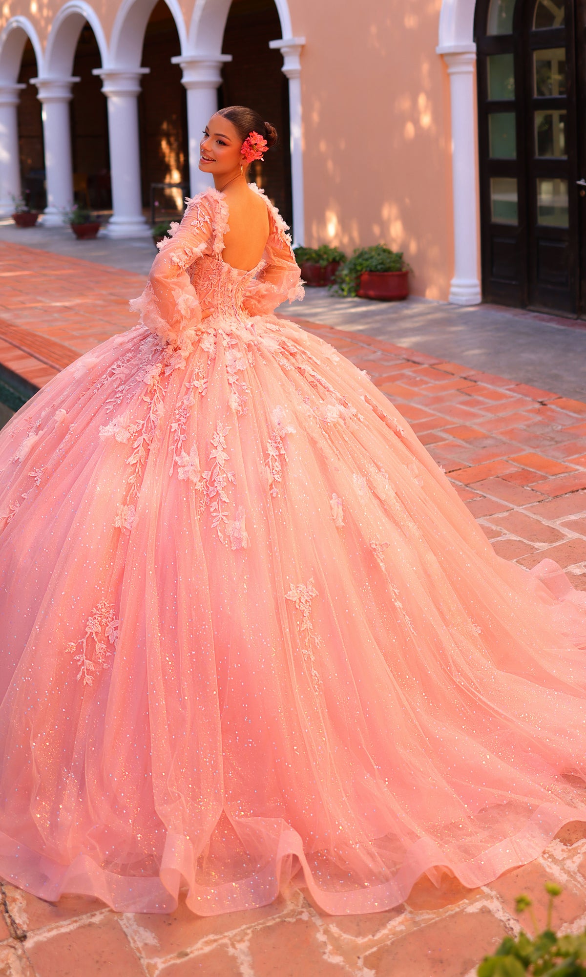 A young woman wearing the Corset Ball Gown Quinceañera Dress 59290 by Amarra stands on a brick walkway outdoors, looking back over her shoulder with arches and potted plants in the background.