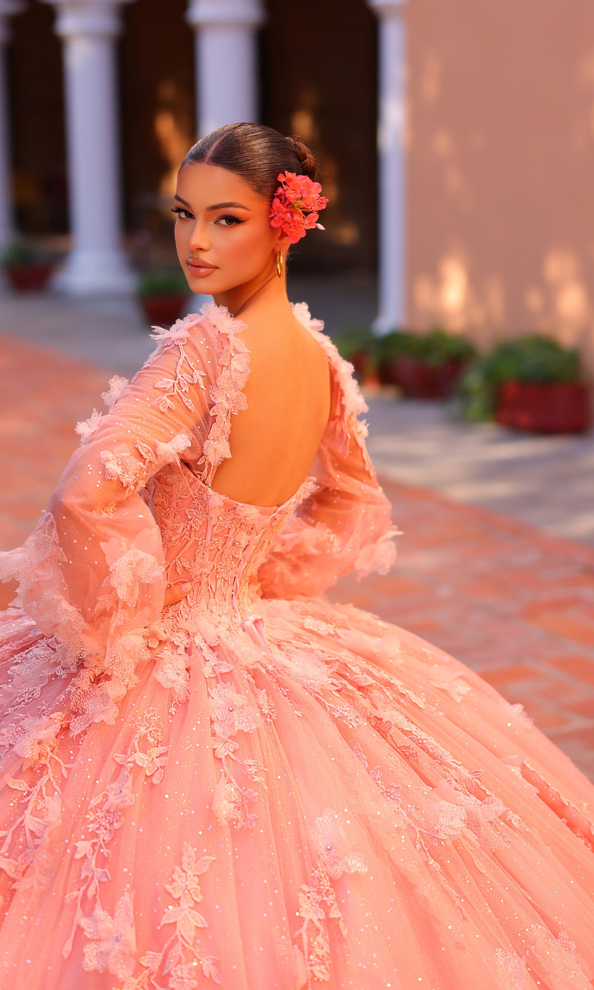 A woman stands outdoors in the Corset Ball Gown Quinceañera Dress 59290 by Amarra, a pink floral gown with a low back and long sleeves. She looks over her shoulder, evoking elegance in a courtyard with a flower in her hair.