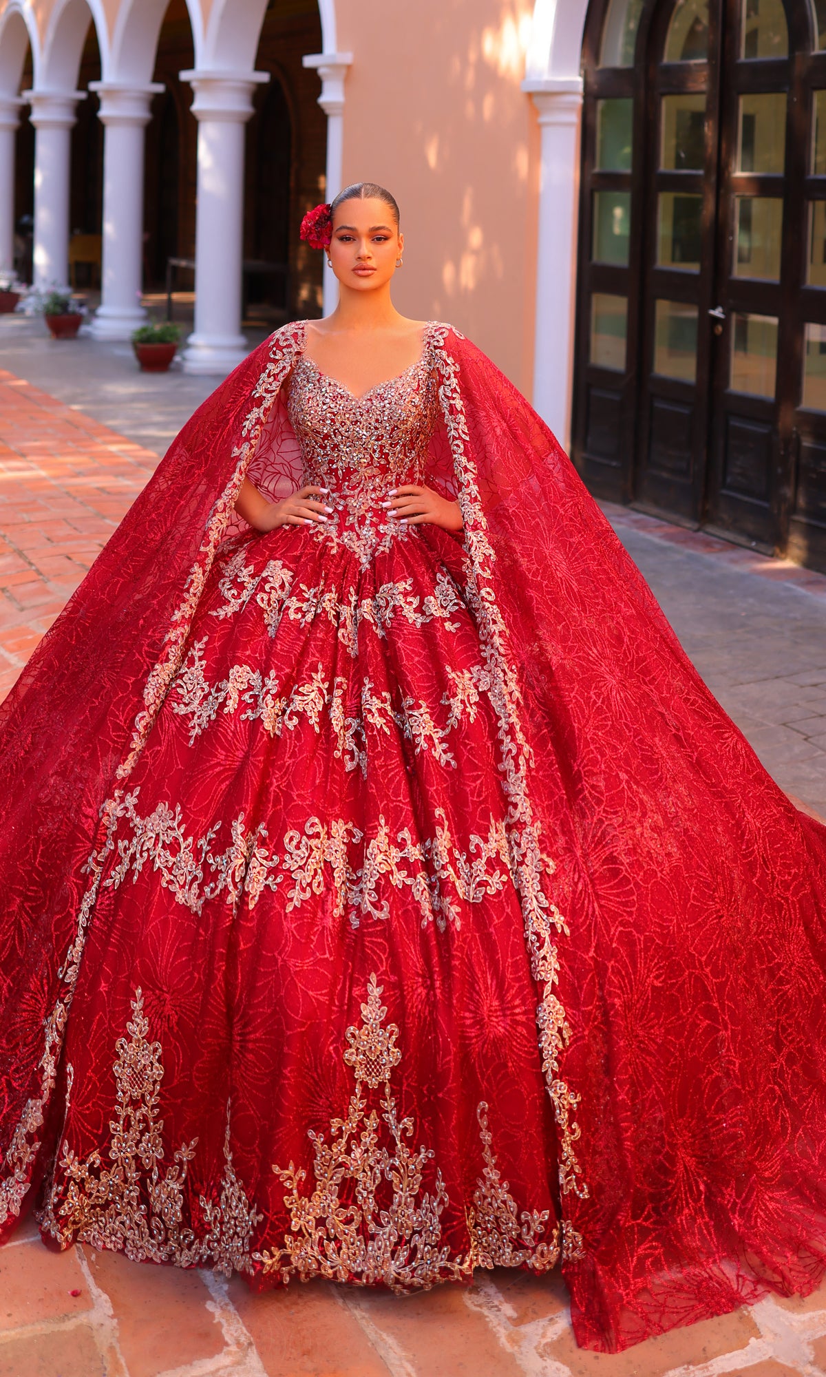 A woman poses elegantly outdoors in the Amarra 54296 Red and Gold Quincea¤era Dress, featuring ornate embroidery and dramatic cape sleeves, before arched doorways.