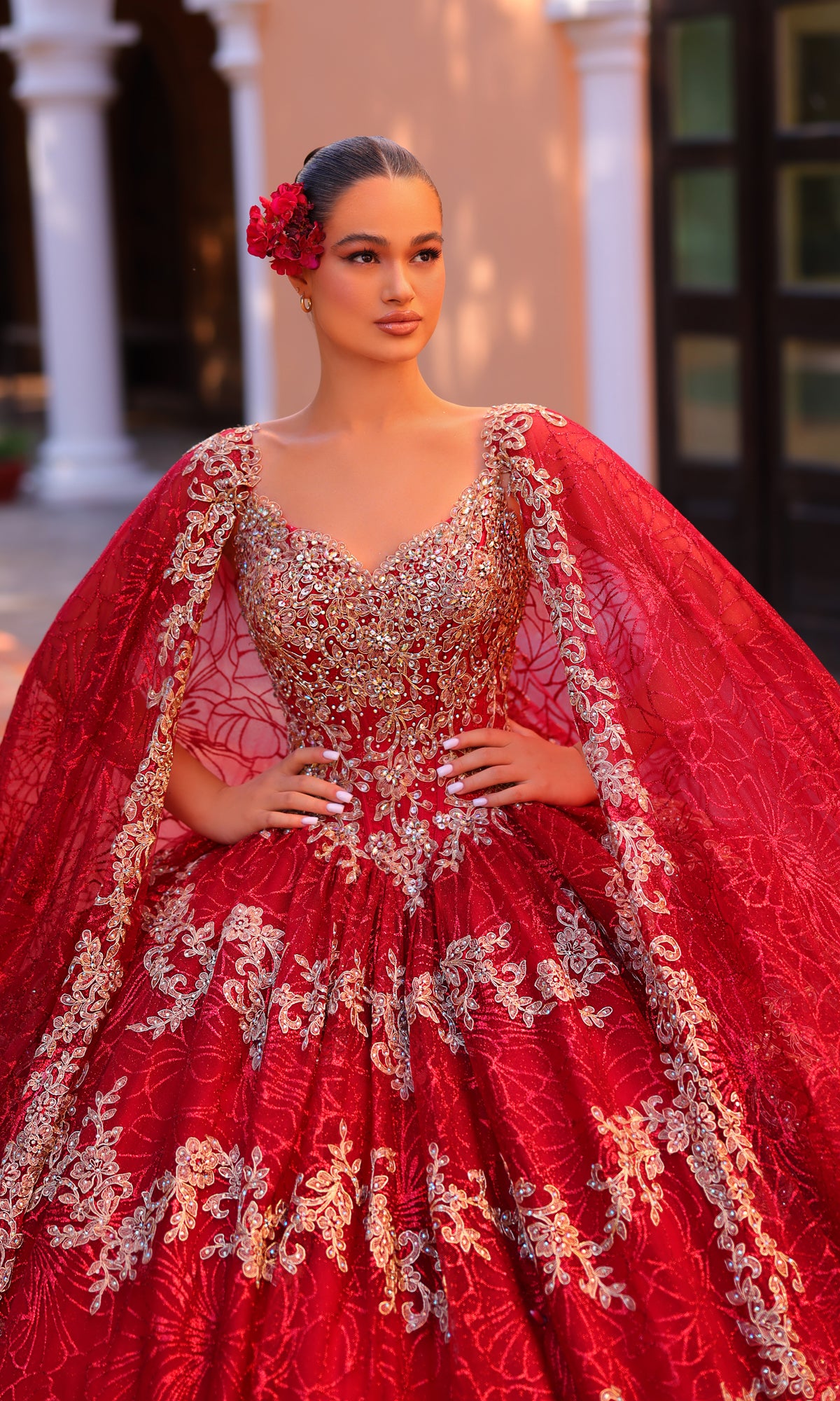 A woman poses confidently, hands on hips, wearing the Amarra 54296 Red and Gold Quincea¤era Dress with cape sleeves and a red flower in her hair.