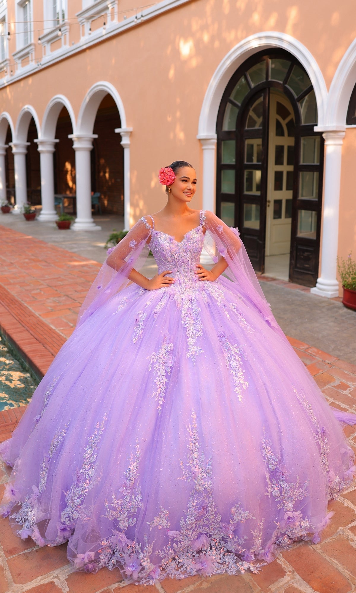 A young woman stands outdoors in front of an arched building, wearing the Quinceanera Dress 54306 by Amarra?a lavender ball gown with floral embroidery and a flower in her hair.