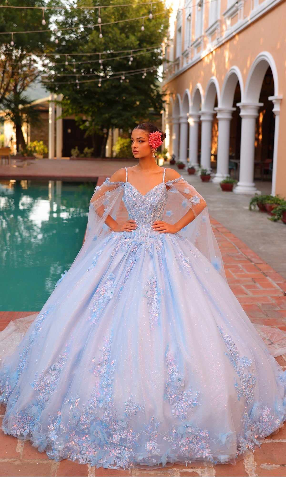 A woman in the Quinceanera Dress 54306 By Amarra, a blue ball gown with a corset bodice and floral embroidery, stands by a pool in a courtyard framed by arched columns and potted plants.